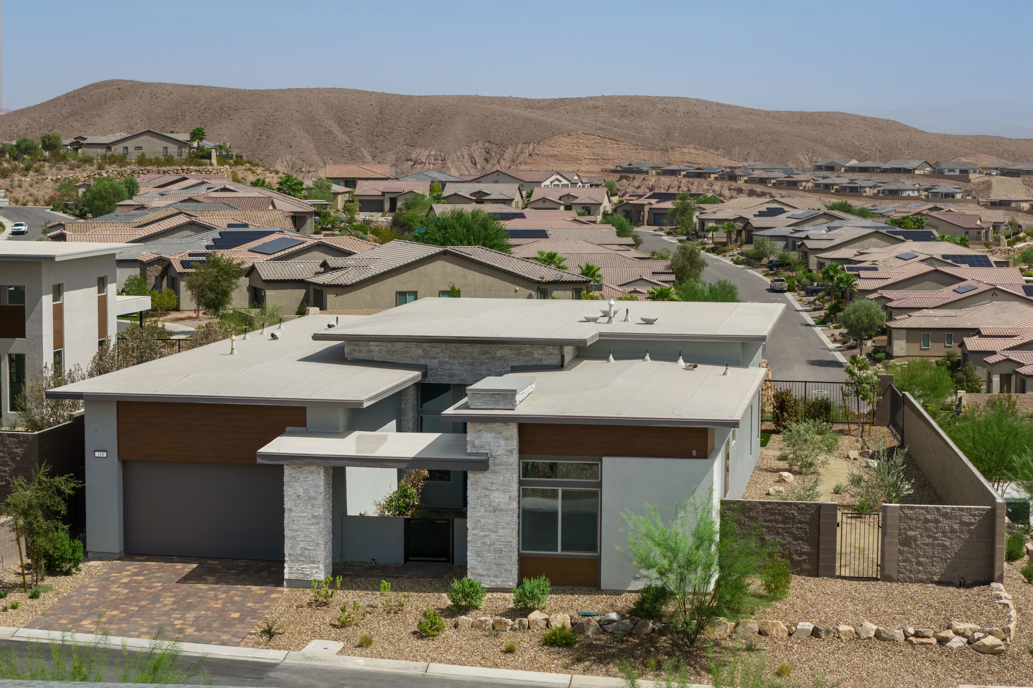 A modern gray house with a flat roof sits in the foreground, surrounded by a sprawling suburban neighborhood with arid hills in the background.