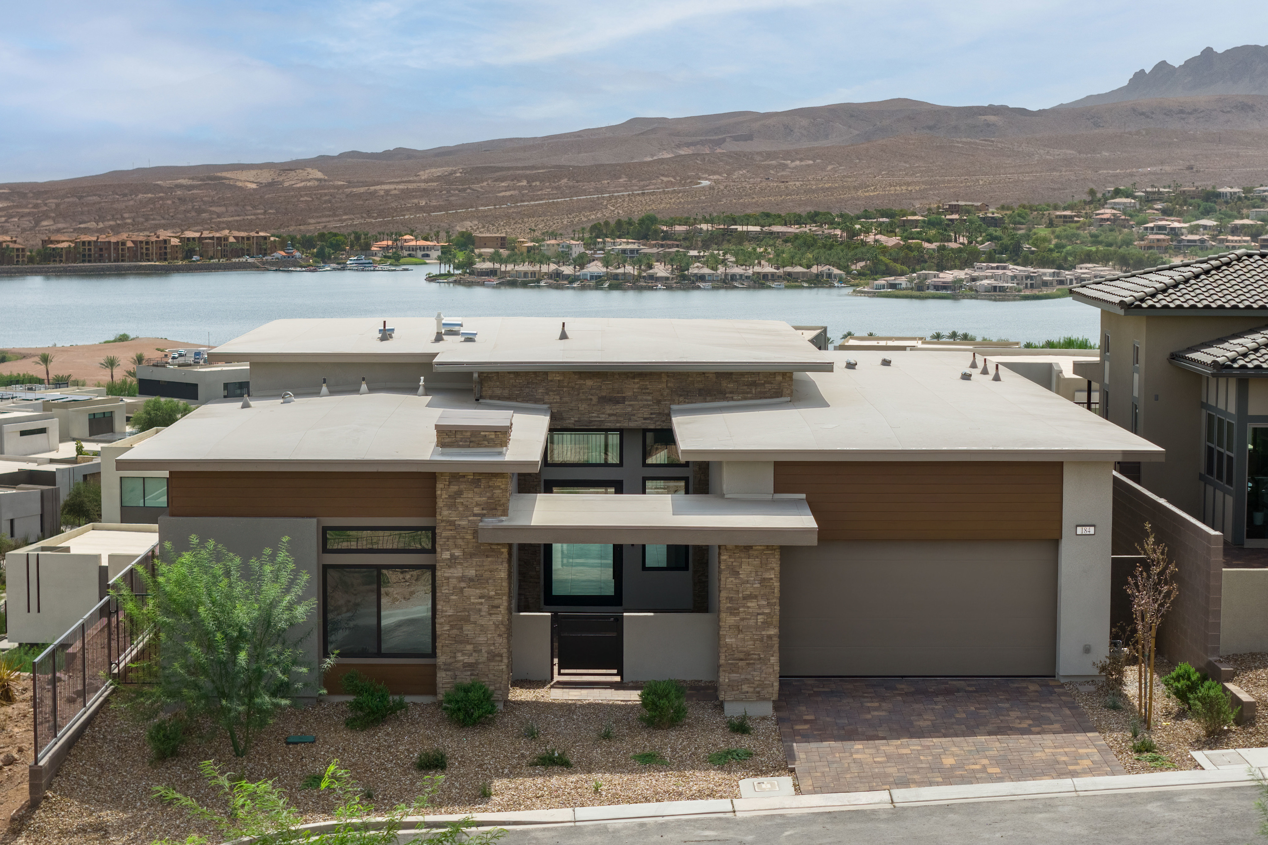 A modern house with stone and wood accents sits in the foreground, overlooking a tranquil lake and distant desert mountains under a hazy sky.