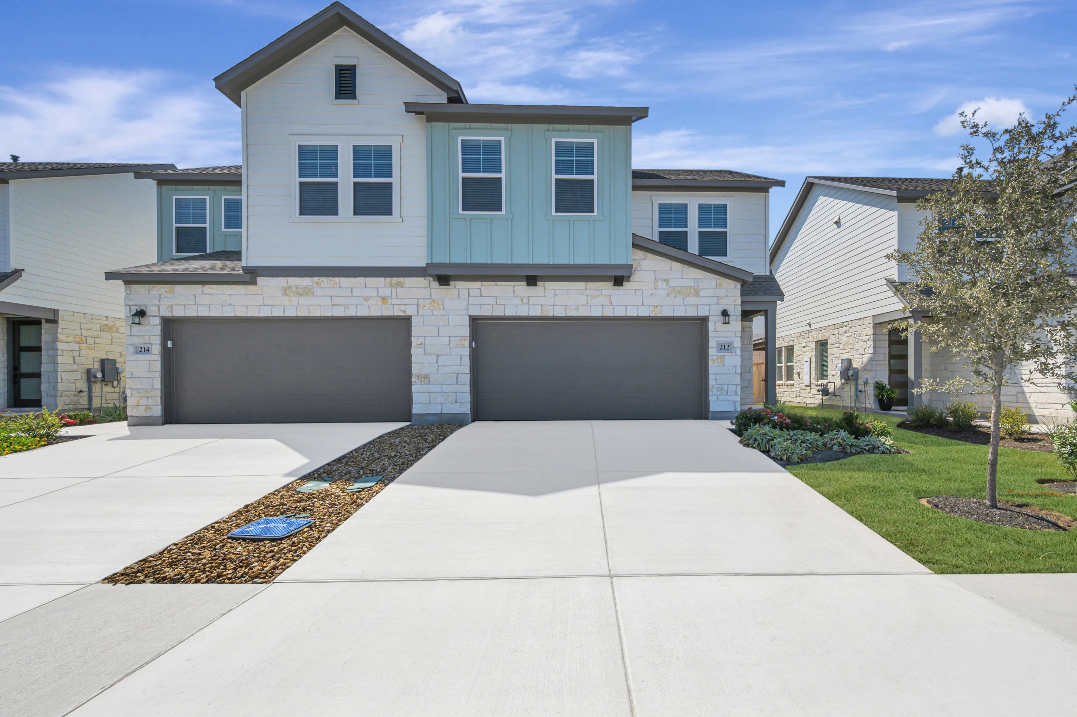 A modern two-story townhouse with a light-colored stone base and blue siding stands on a bright concrete driveway, with a small tree and manicured lawn to the right, under a blue sky with scattered clouds.