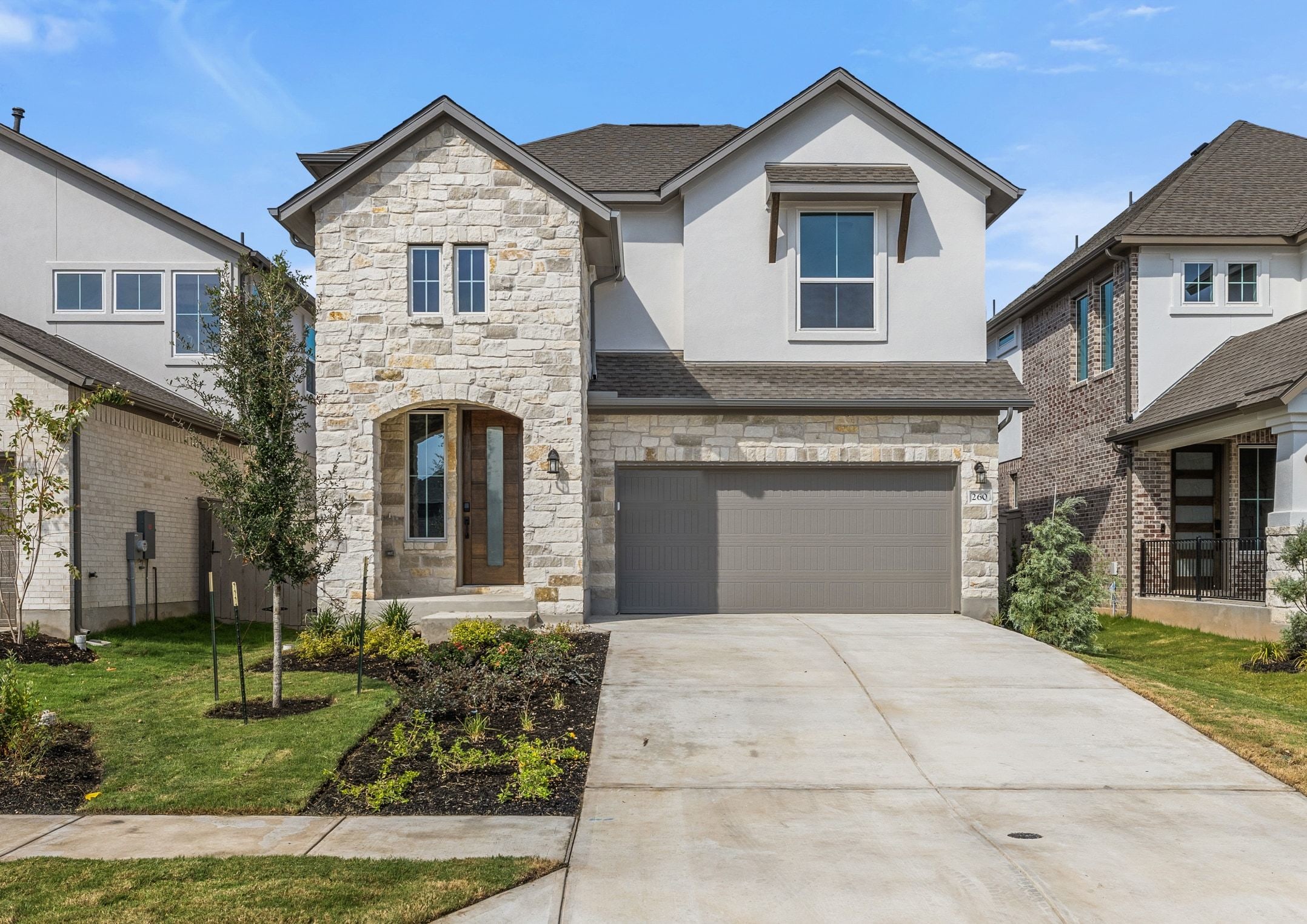 A modern stone and stucco house with a gray garage door sits on a concrete driveway in front of a clear blue sky, with manicured lawns and landscaping in the foreground and neighboring houses visible in the background.