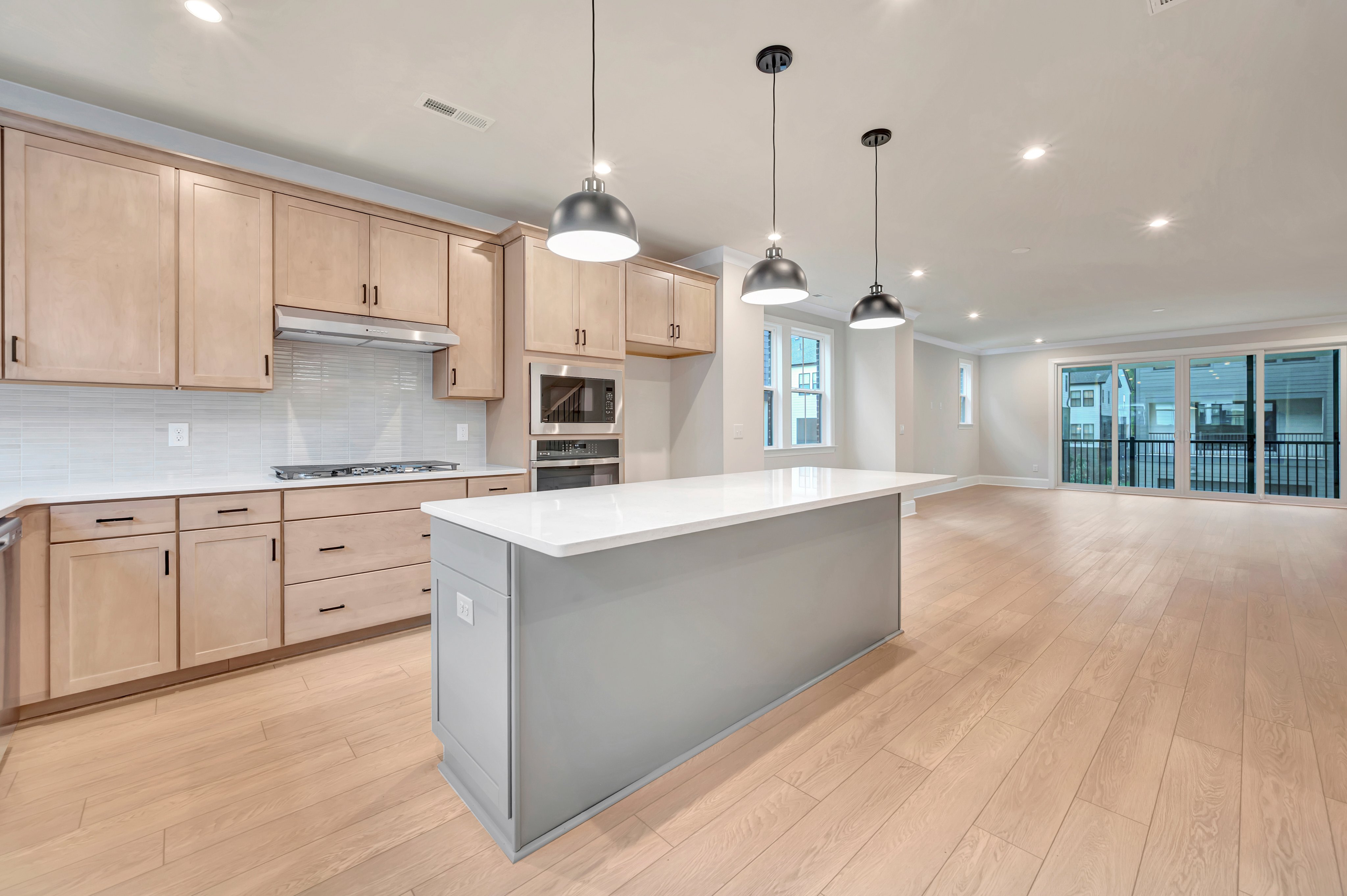 A modern kitchen with a light gray island in the foreground opens into a spacious living area with large glass doors in the background.