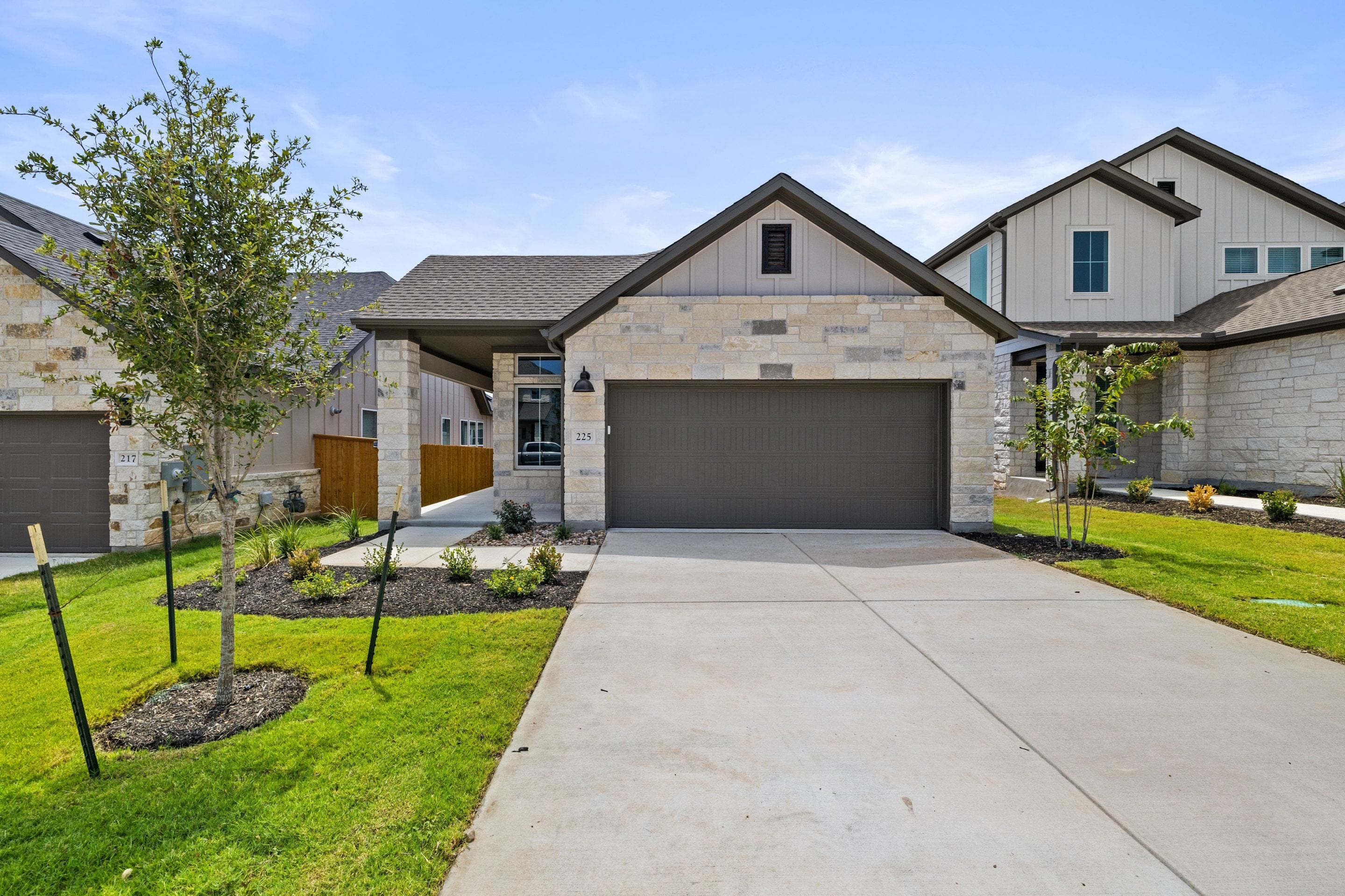 A modern stone-clad house with a dark garage door sits on a concrete driveway in the foreground, while other similar houses and a clear blue sky form the background.