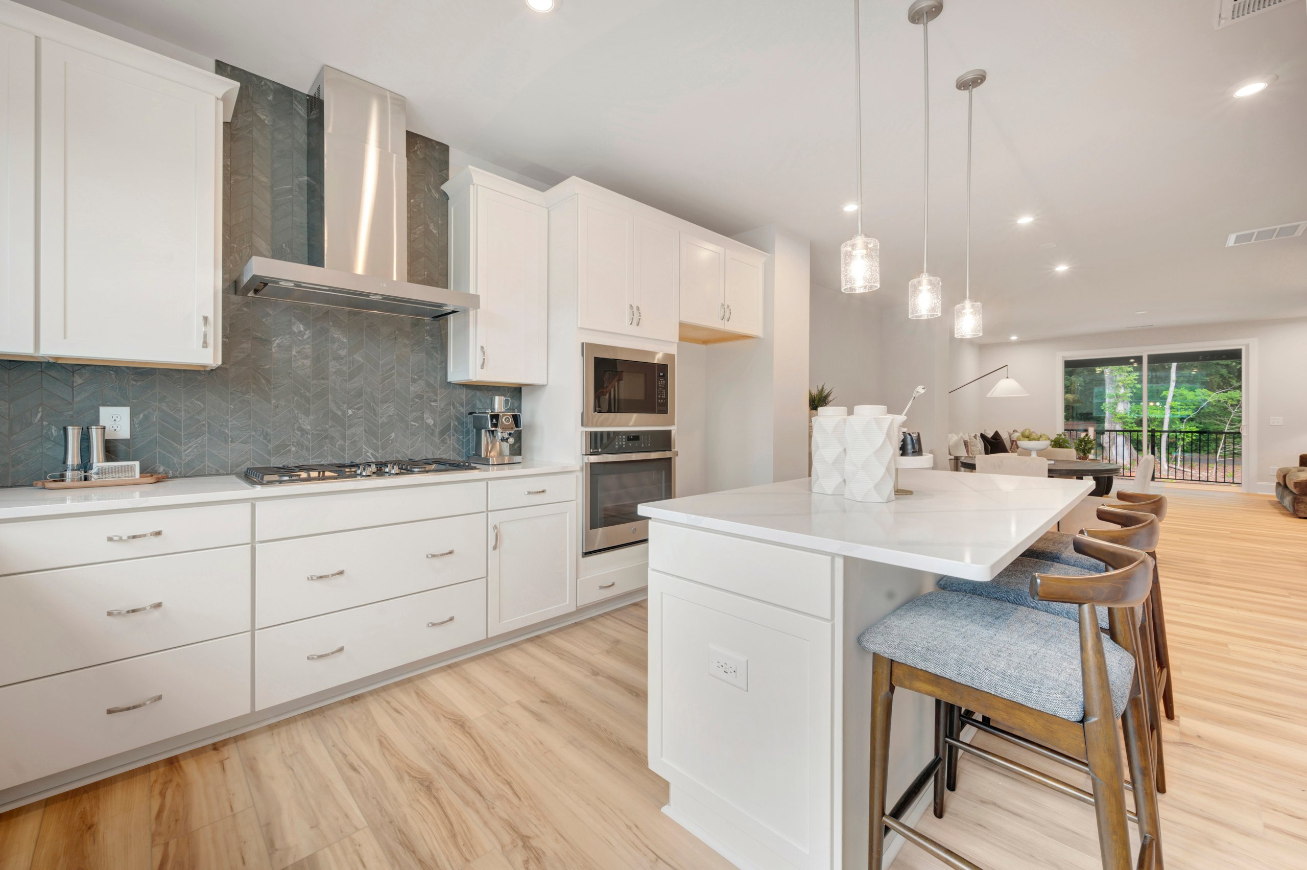A modern white kitchen with a marble island and bar stools in the foreground is connected to a bright living area with large windows in the background.