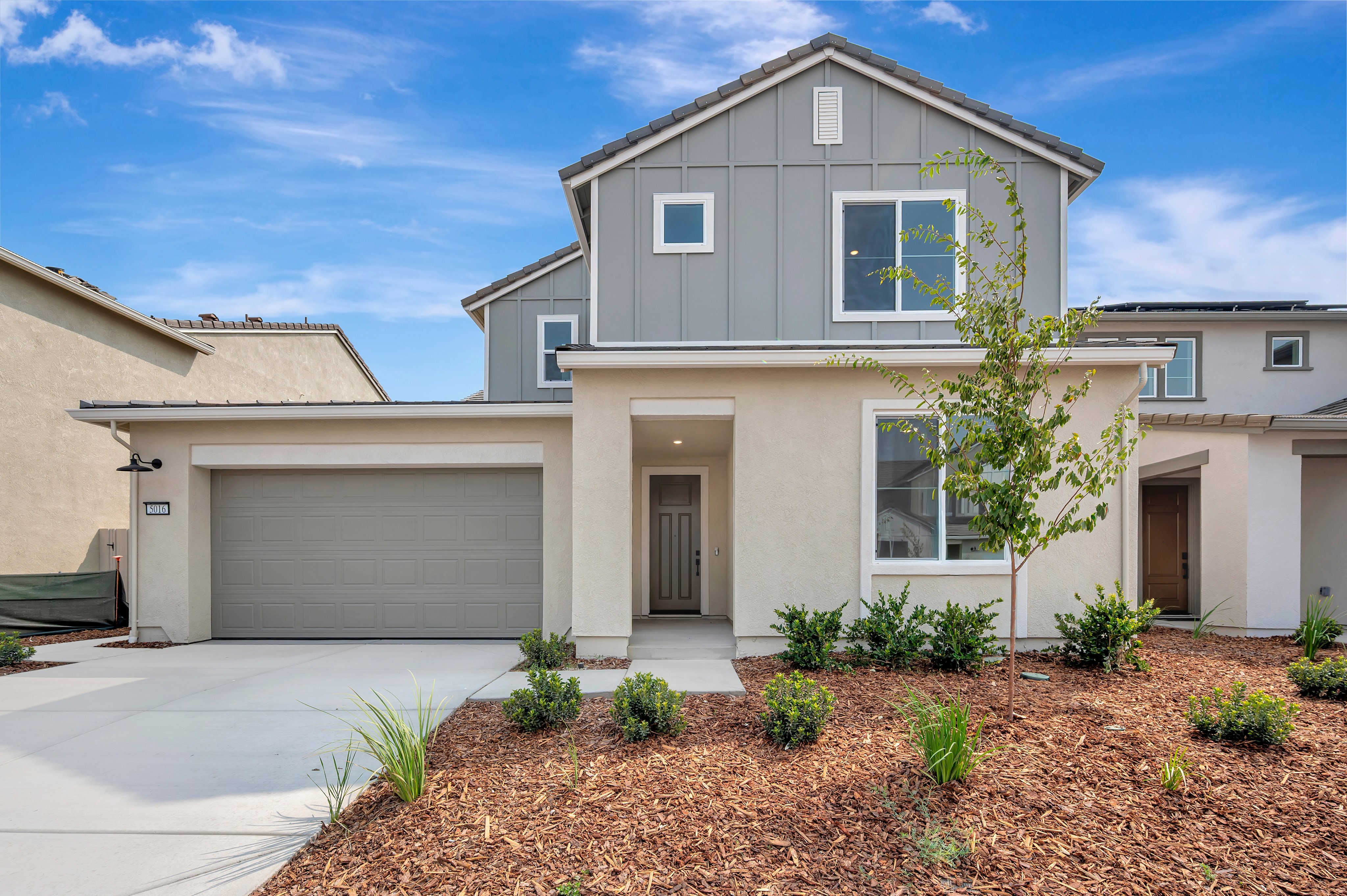 A modern two-story house with a beige and gray exterior and a manicured lawn sits under a partly cloudy blue sky.