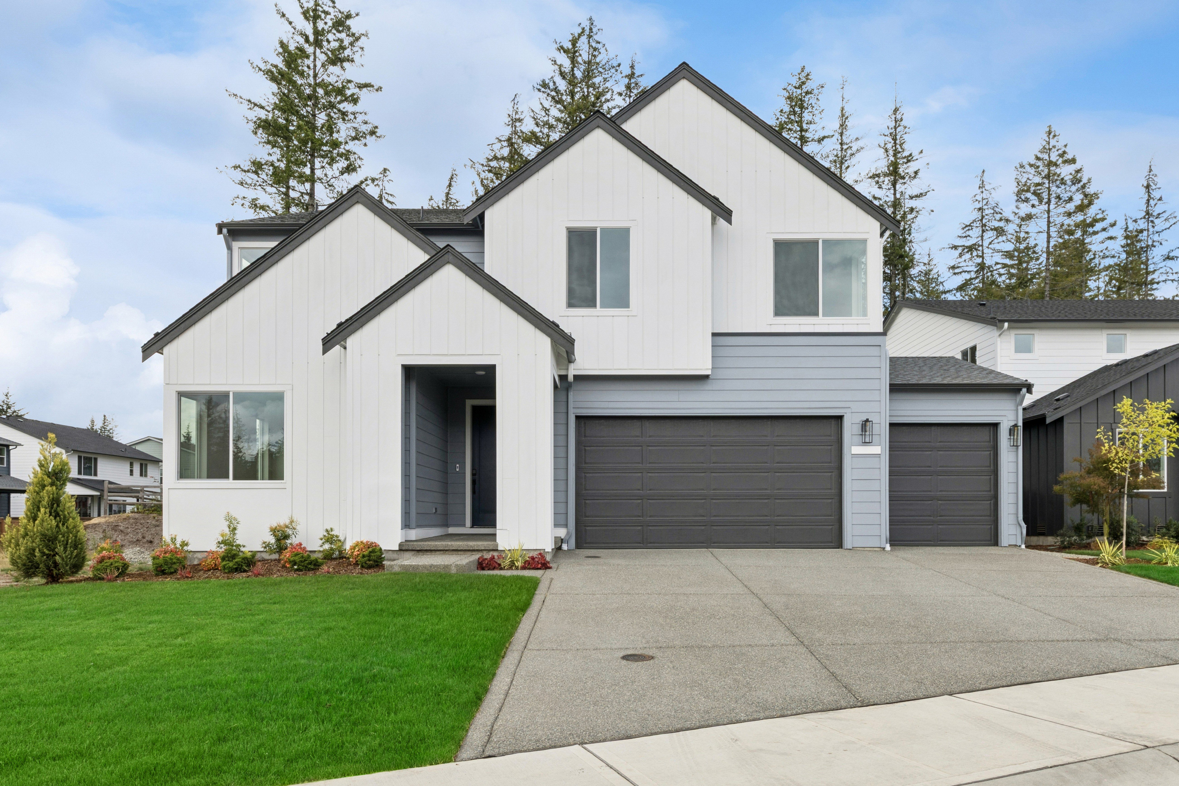 A modern white and gray house with a manicured lawn in the foreground is set against a backdrop of evergreen trees and a blue sky.
