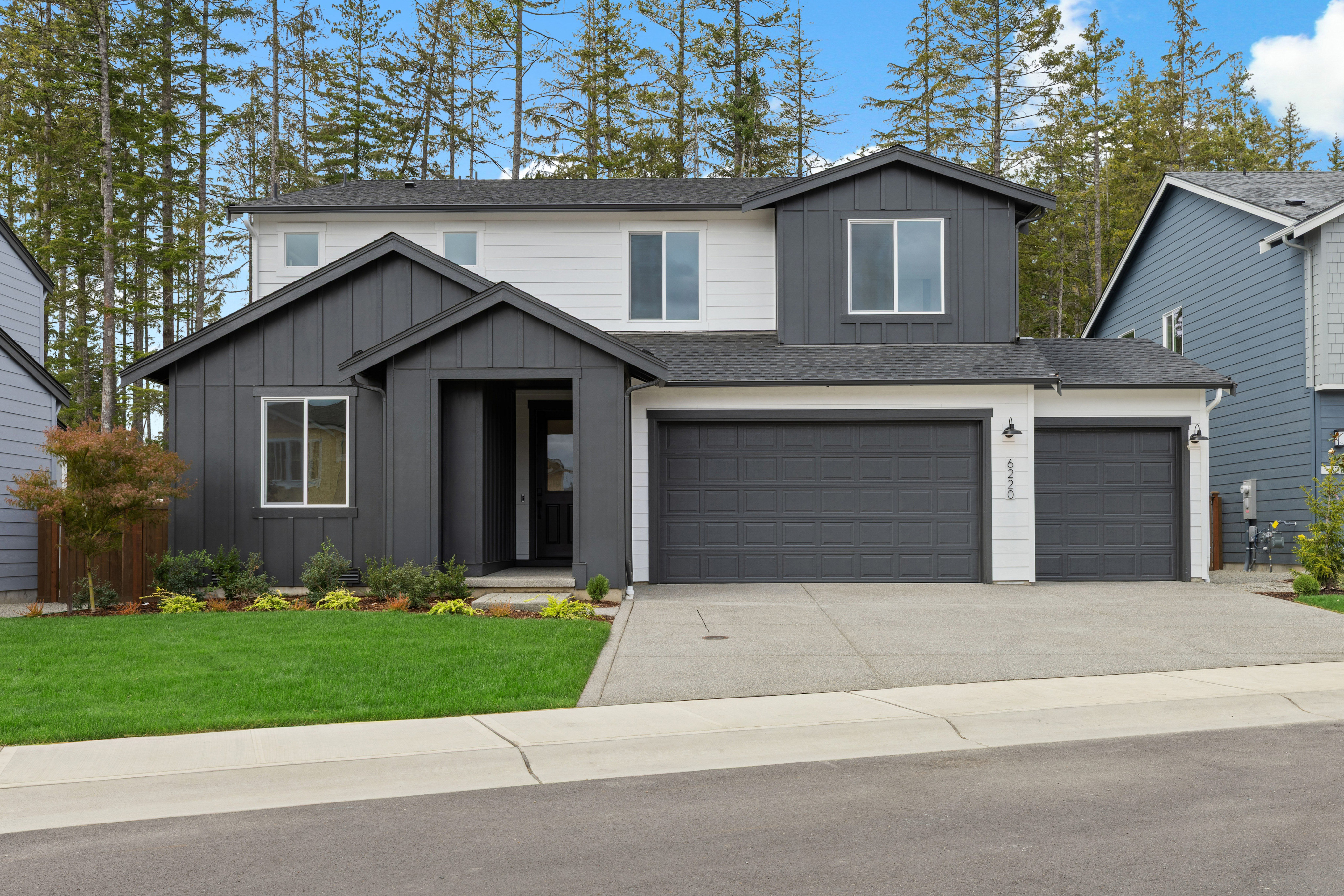 A modern two-story house with a dark gray and white exterior and a two-car garage sits on a manicured lawn, backed by a dense forest under a bright blue sky.