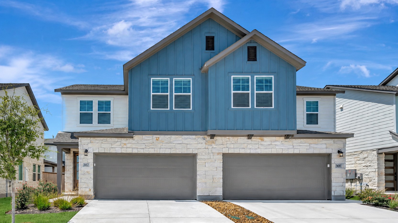 A concrete driveway with a red and white striped curb leads to a row of modern townhouses with blue and white siding and stone accents, set against a bright blue sky with scattered clouds.