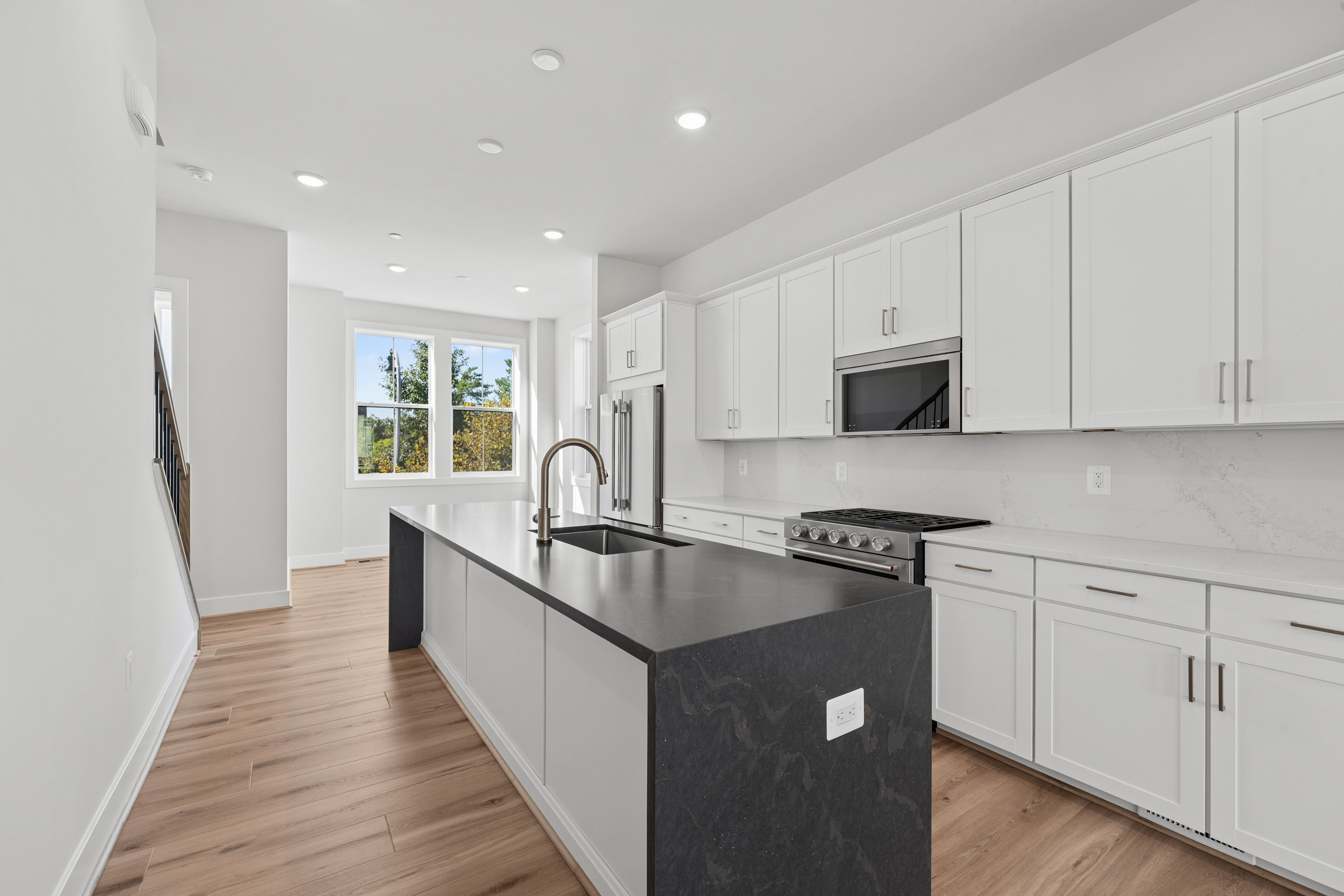 A modern kitchen with a dark island in the foreground and white cabinets and a window with a view of trees in the background.