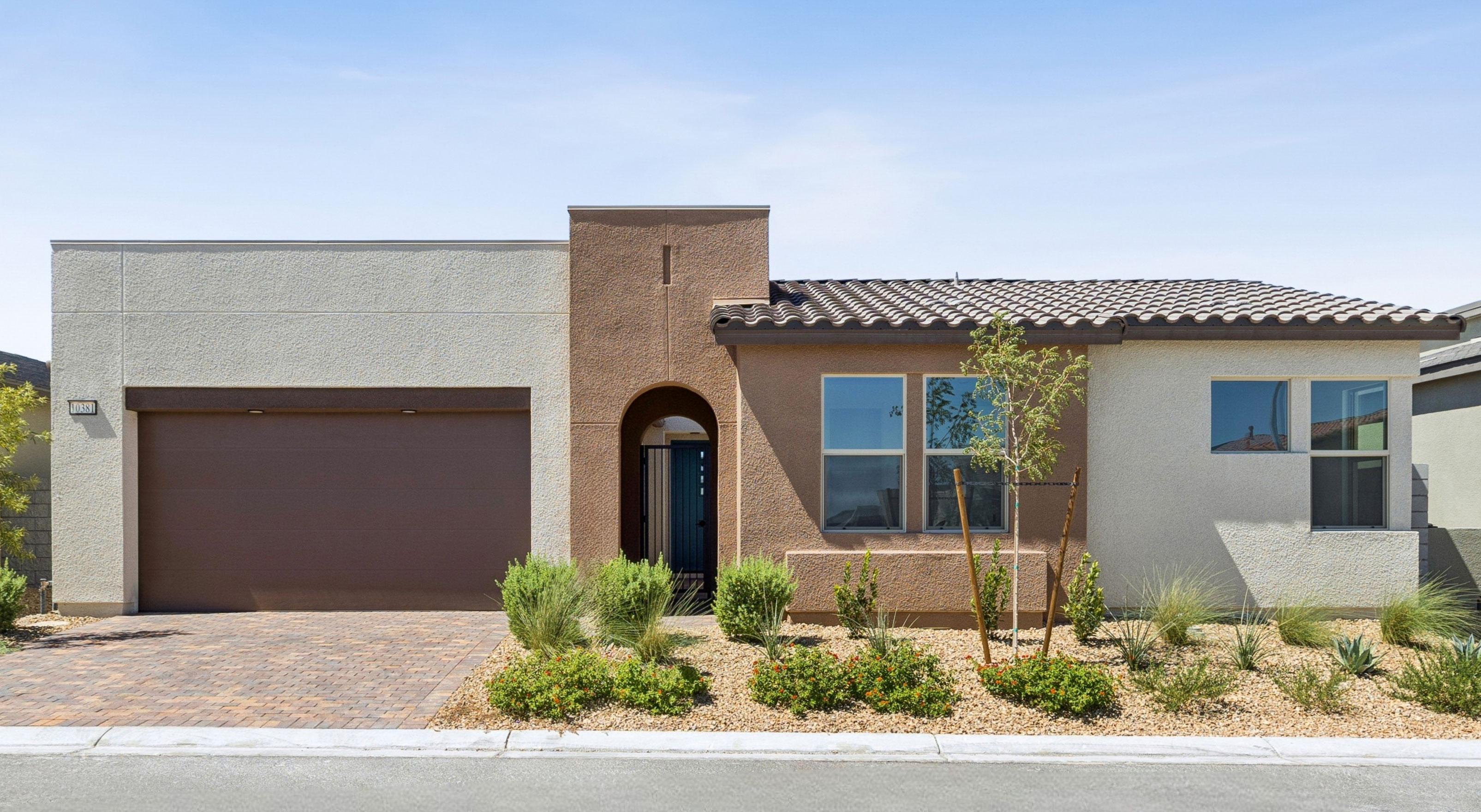 A modern stucco house with a brown garage door and arched entryway sits in the foreground, with a clear blue sky in the background.