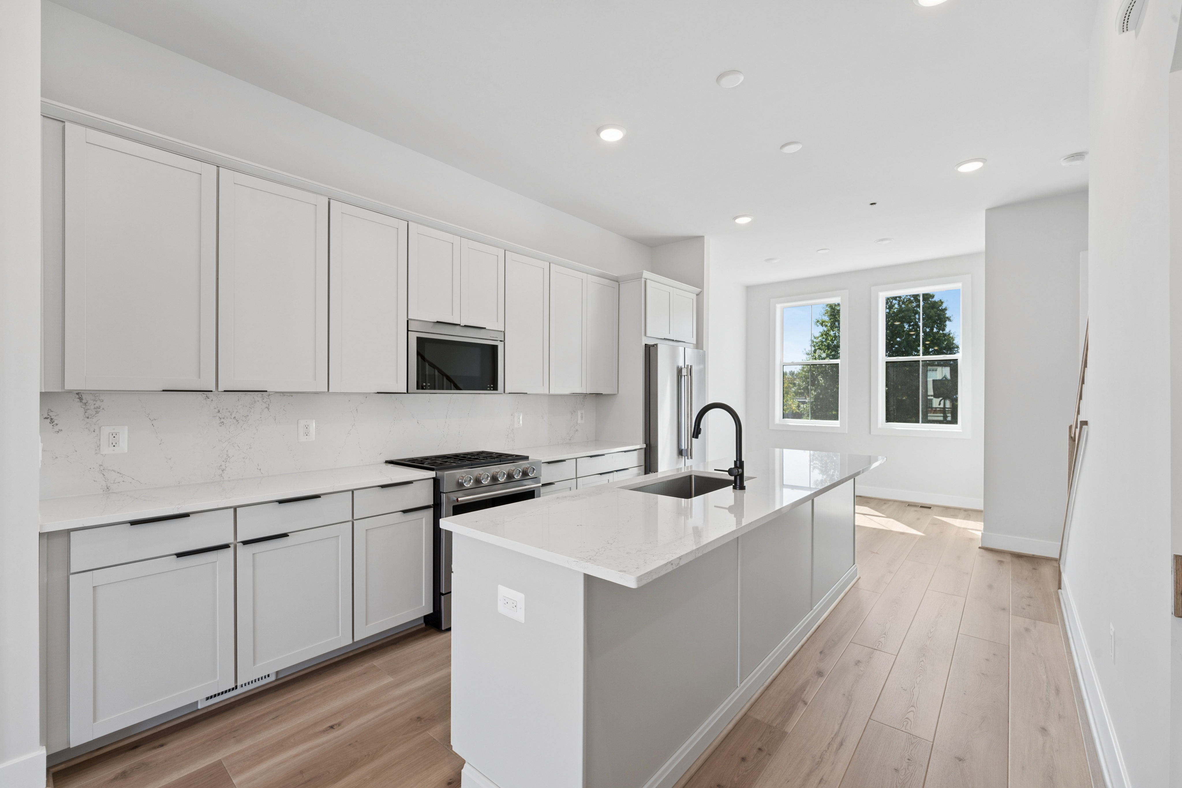 A modern white kitchen with a large island in the foreground and a bright living area with windows in the background.