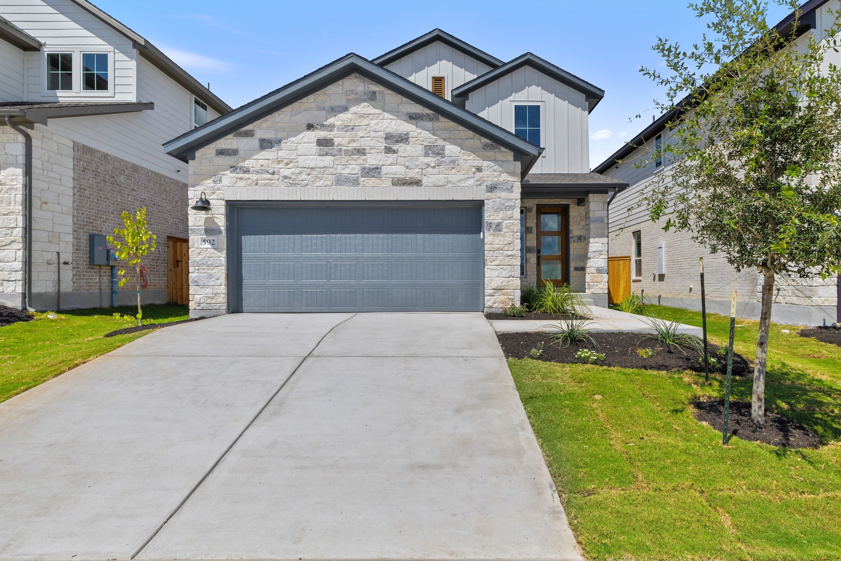 A modern stone-clad house with a gray garage door sits on a concrete driveway, with manicured lawns and trees in the foreground and neighboring houses in the background.