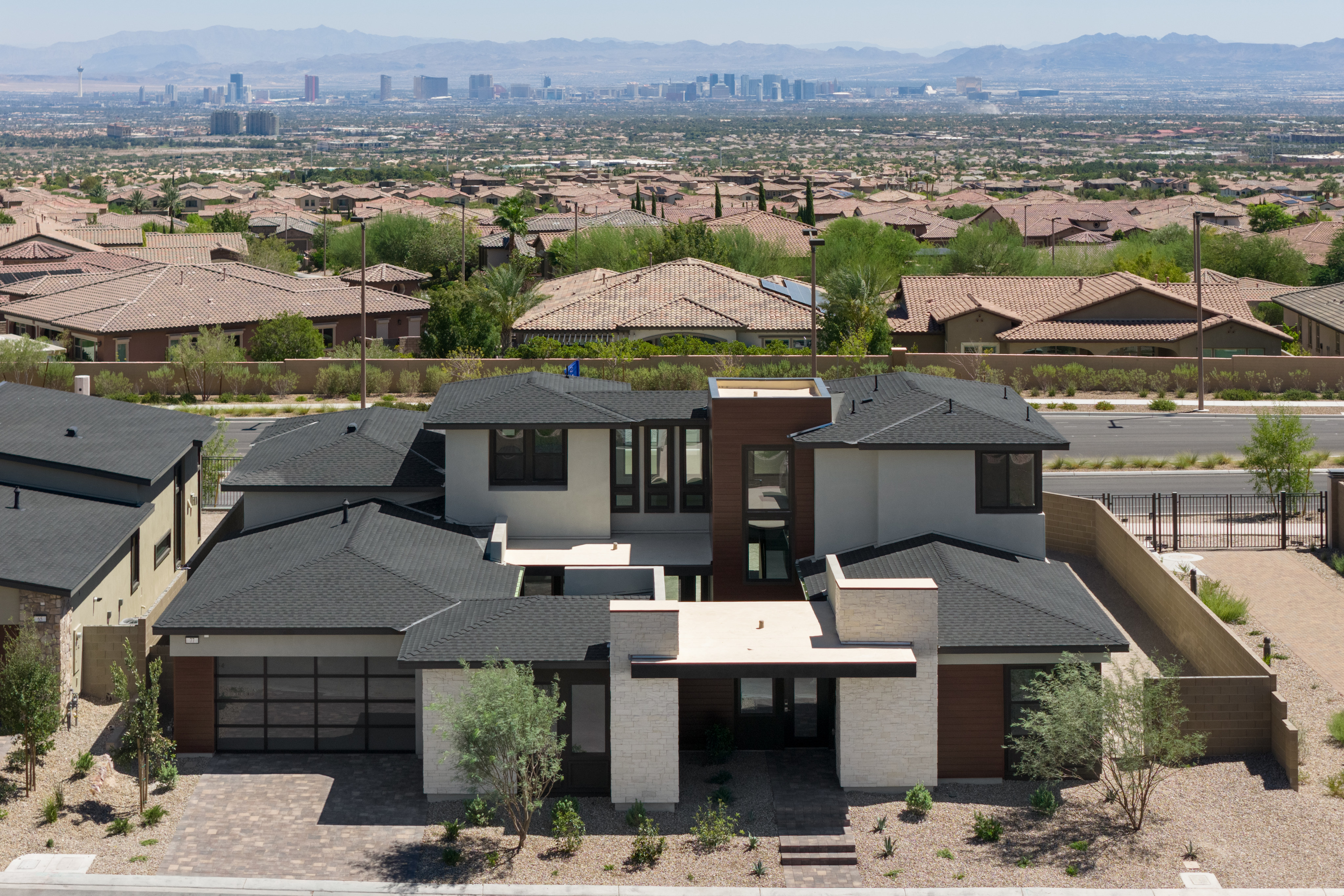 A modern, multi-story house with a dark roof and light-colored walls sits in the foreground, while a sprawling suburban neighborhood with a distant city skyline stretches across the background.