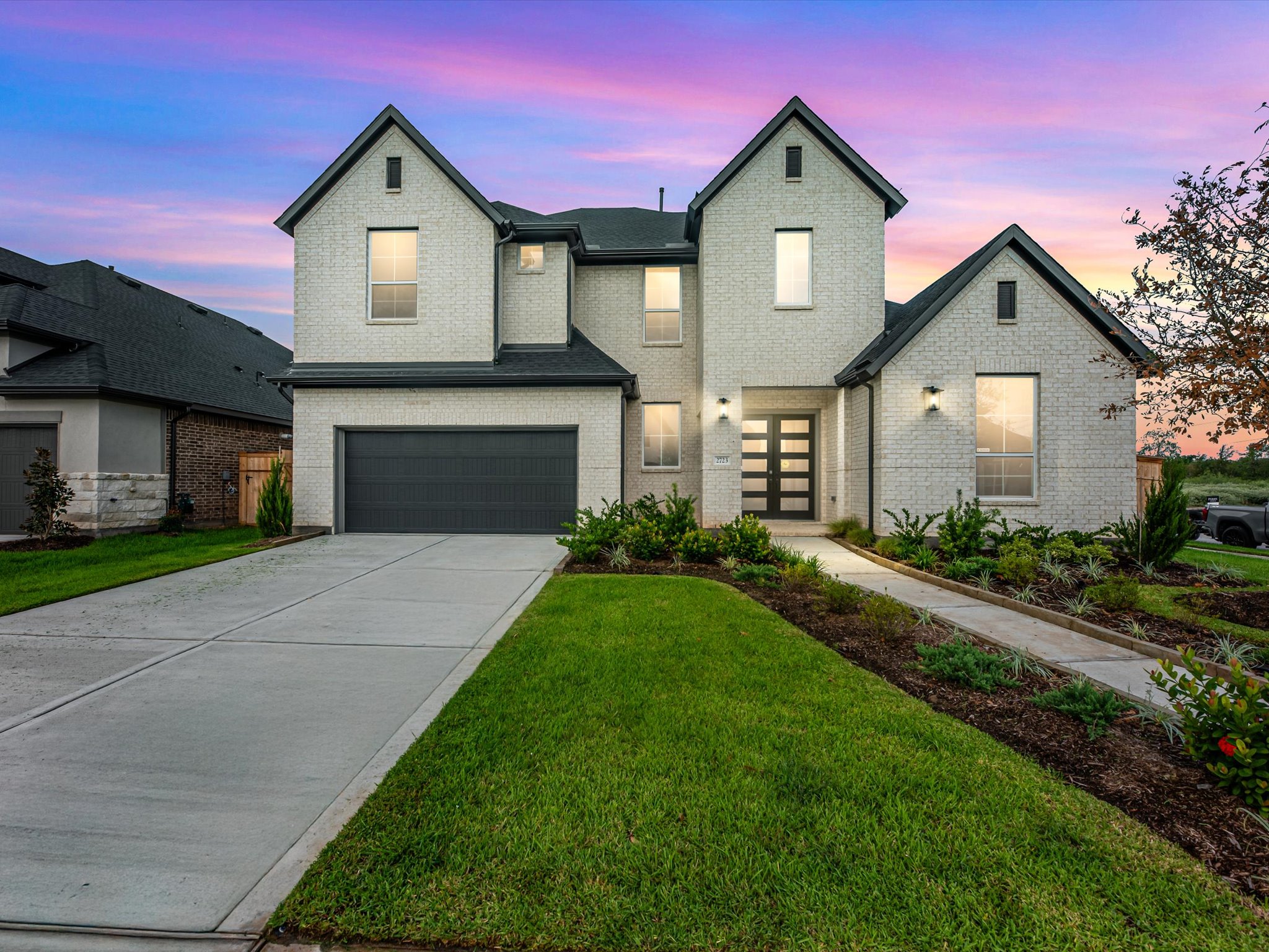 A large white brick house with a grey garage and driveway sits behind a green lawn under a vibrant purple and blue sunset sky.