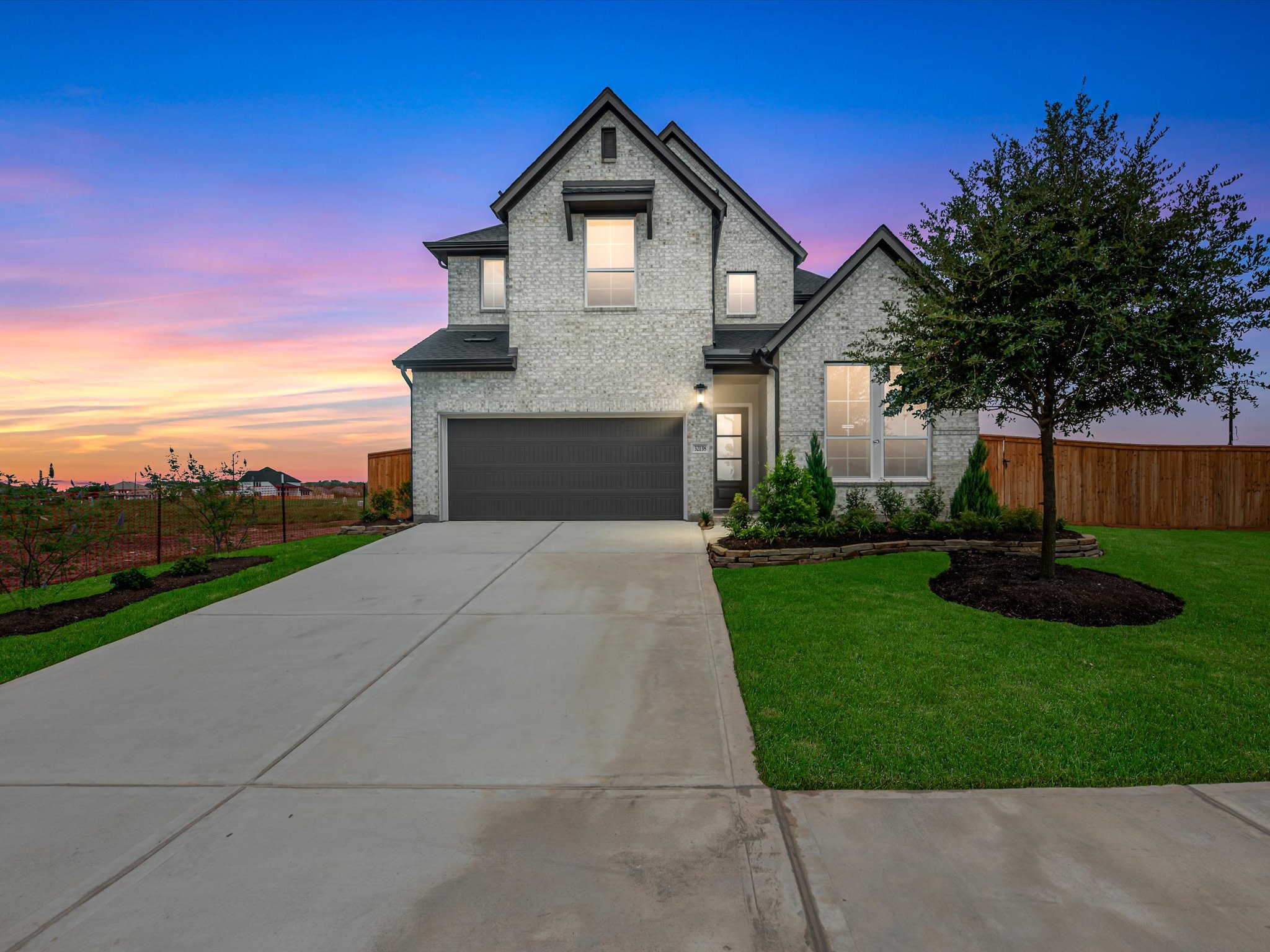 A modern grey brick house with a concrete driveway and green lawn sits under a vibrant sunset sky.