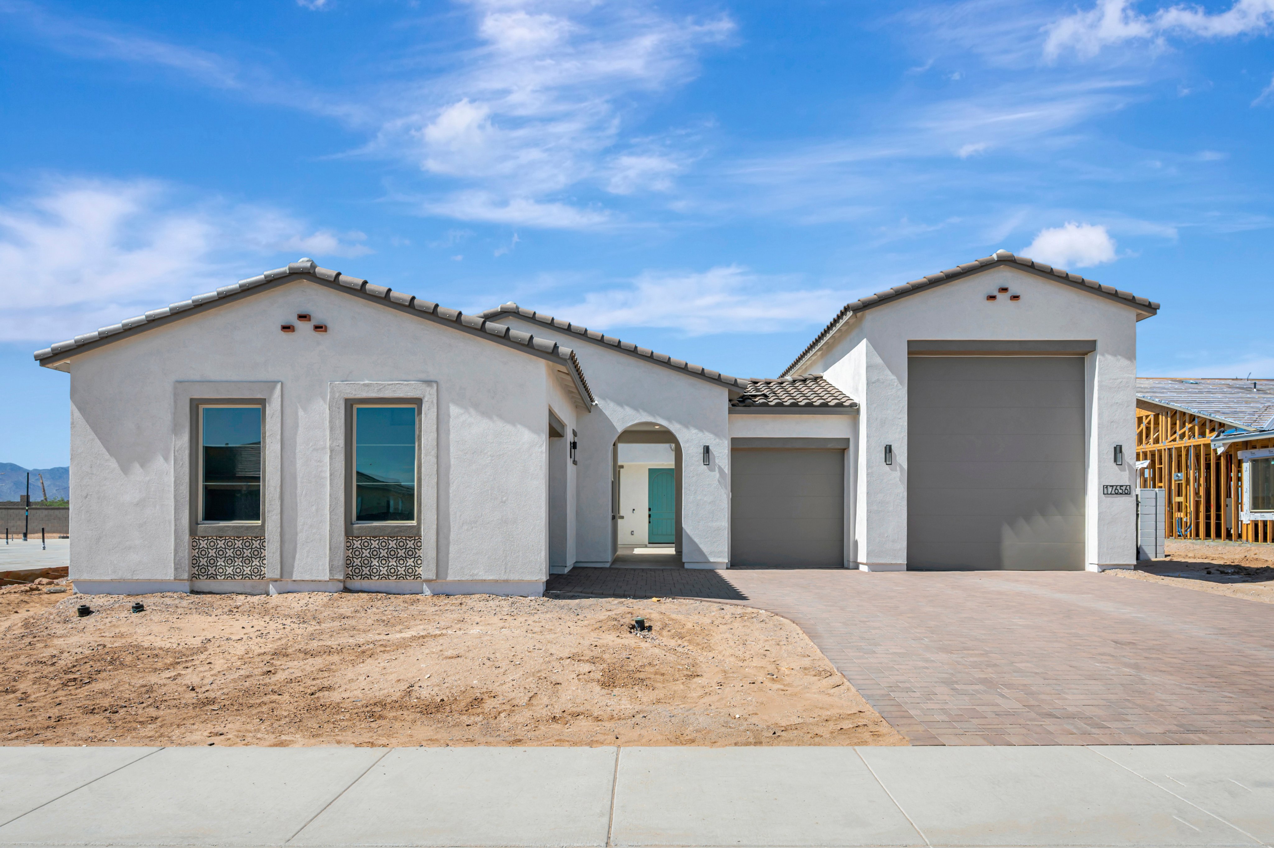 A modern white single-story house with a grey garage stands behind a dirt yard and paved driveway under a bright blue sky with wispy clouds.