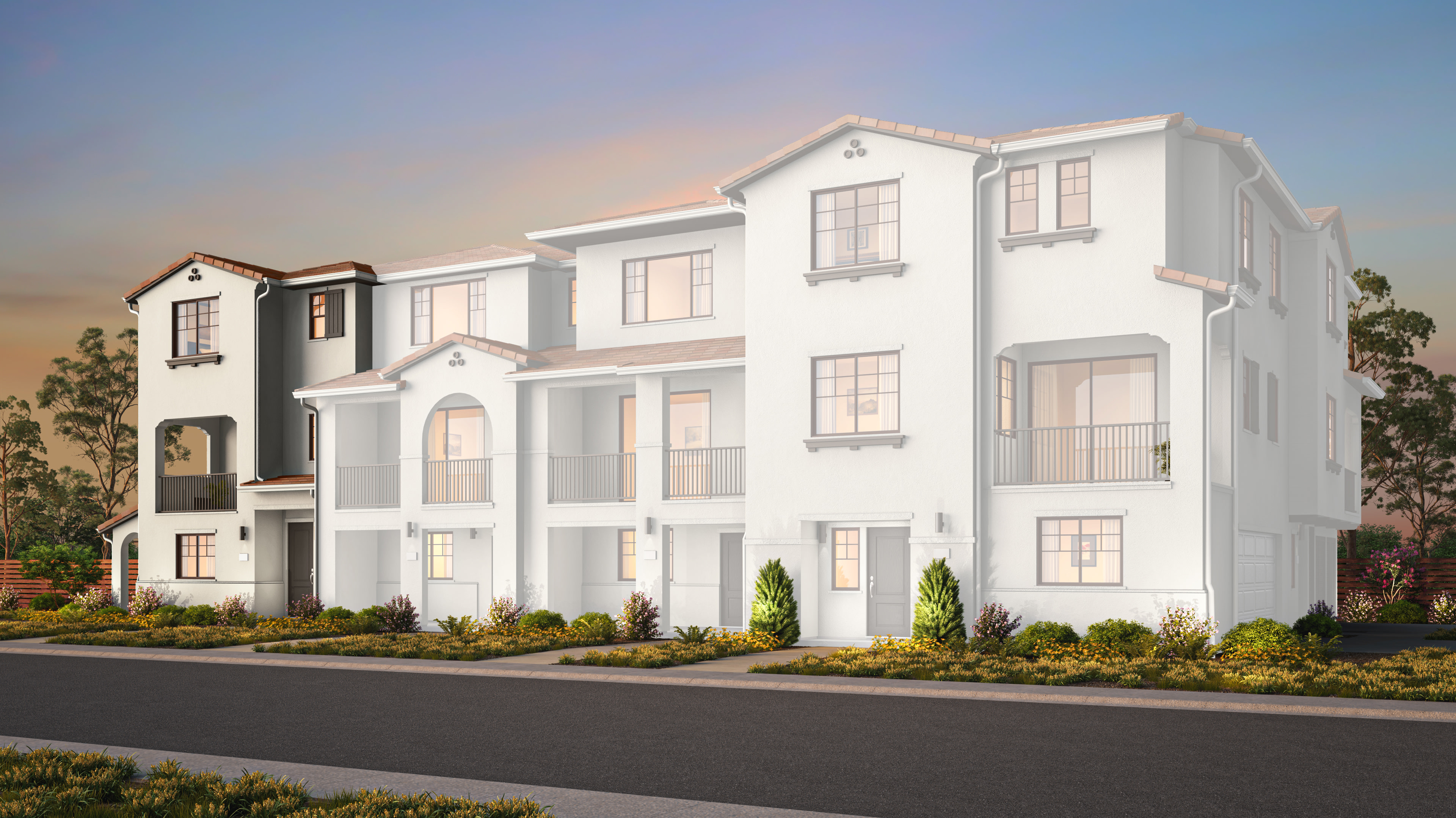 A modern white multi-unit apartment complex stands behind a paved road and manicured landscaping under a soft sunset sky.