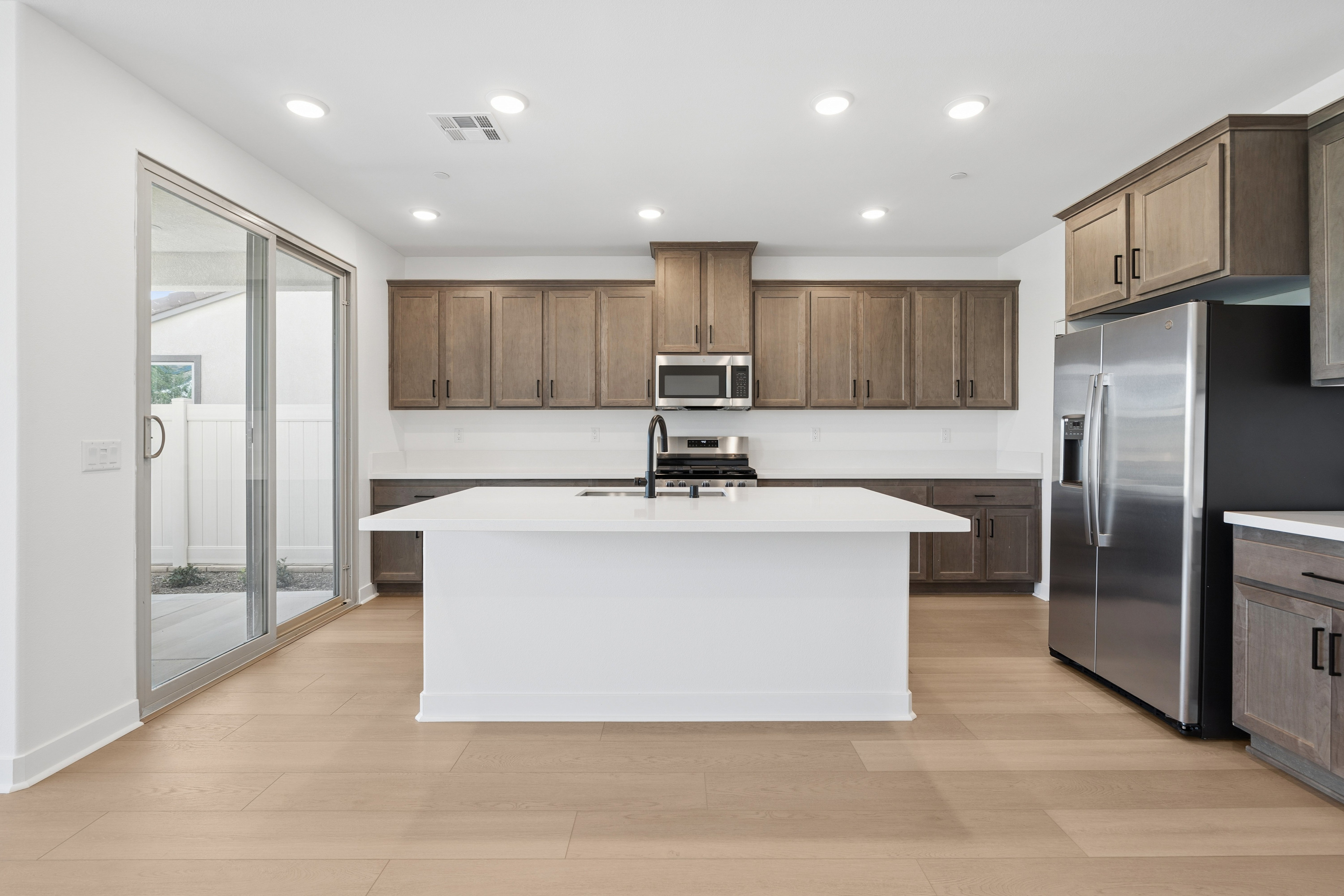 A large white kitchen island sits in the foreground of a modern kitchen featuring grey cabinetry, stainless steel appliances, and a sliding glass door leading outside.