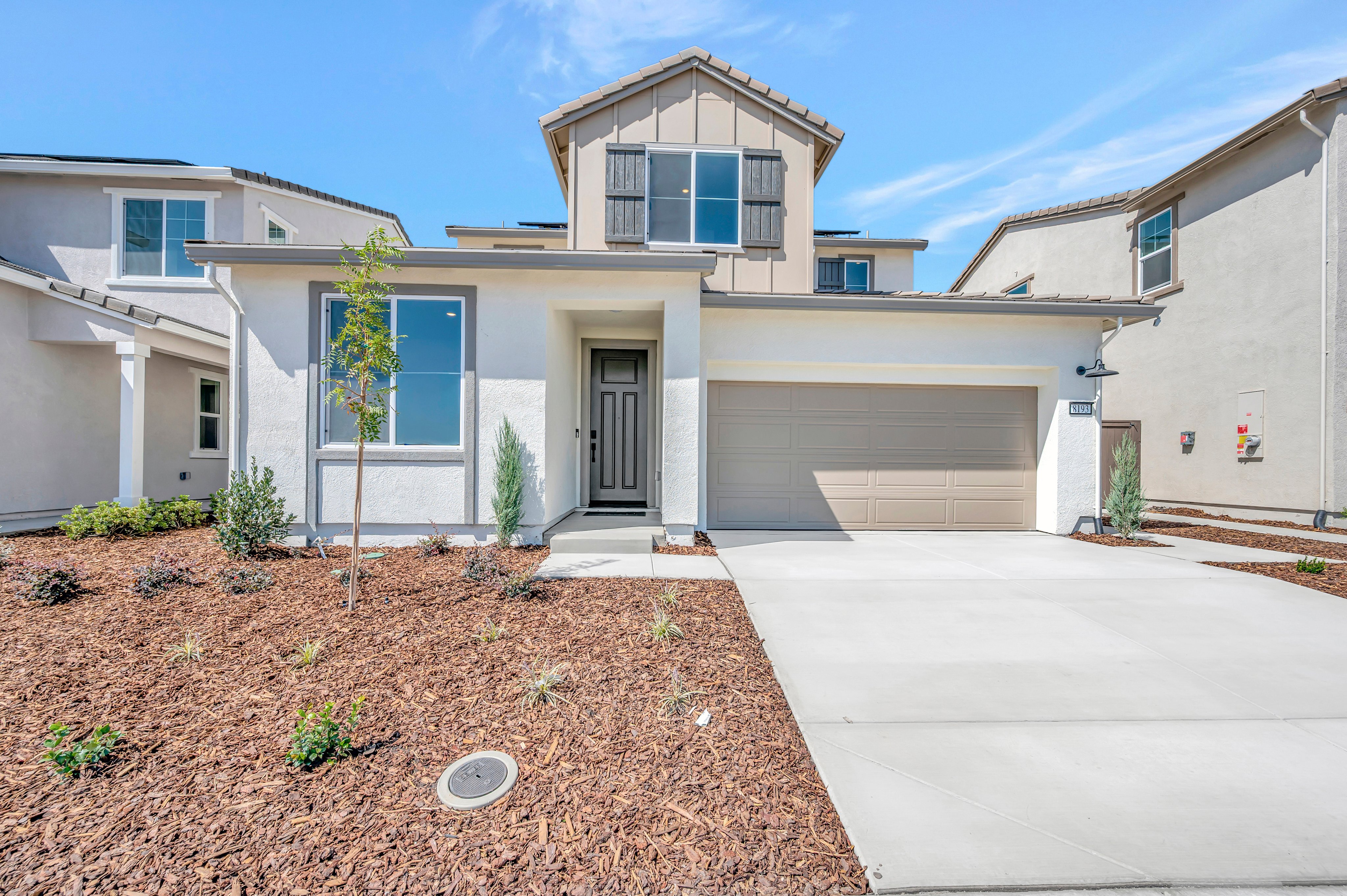 A modern two-story house with a tan garage and white exterior stands behind a mulched yard and concrete driveway under a clear blue sky.