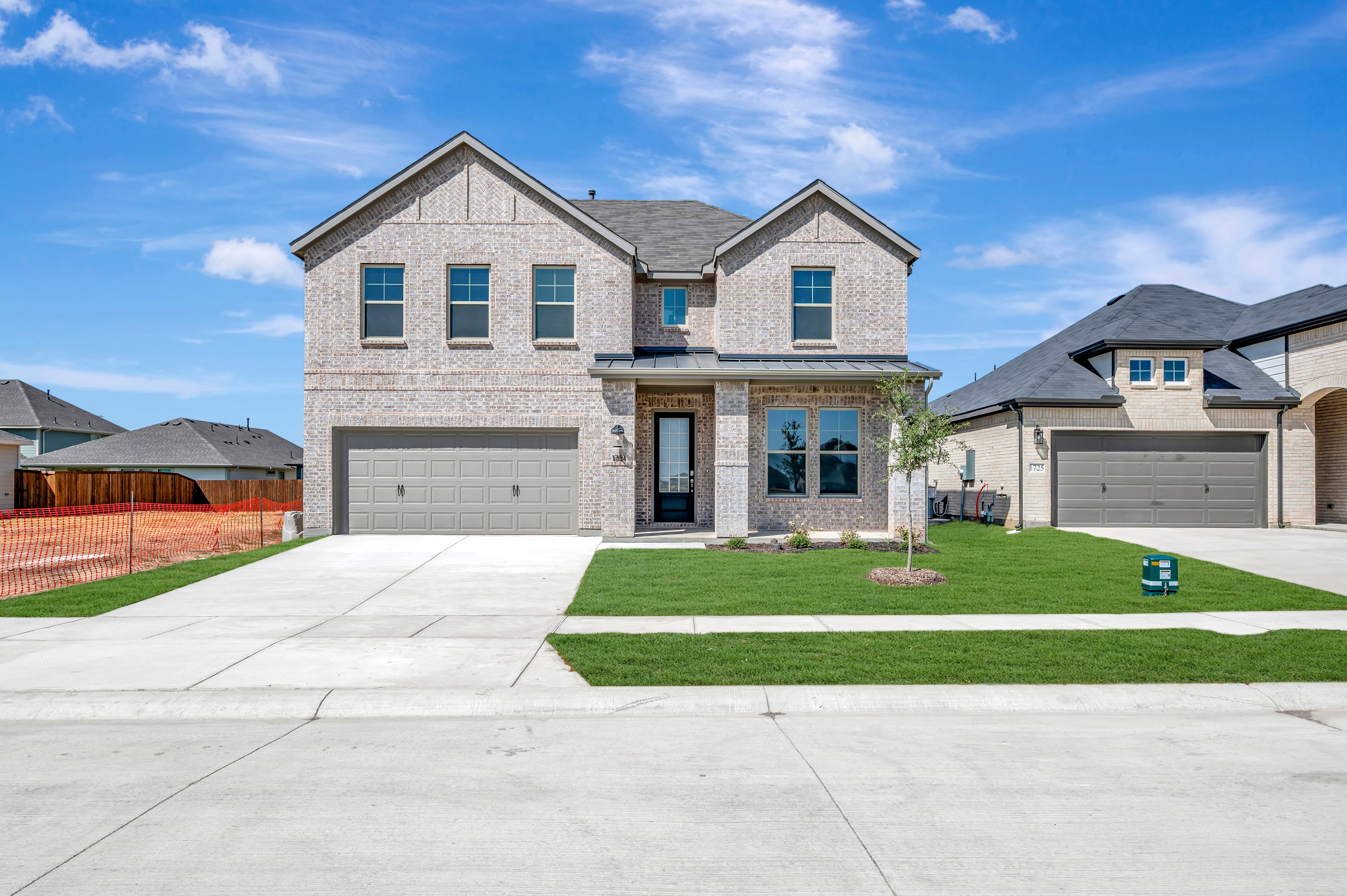 A two-story brick house with a concrete driveway and green lawn sits under a bright blue sky with wispy clouds.