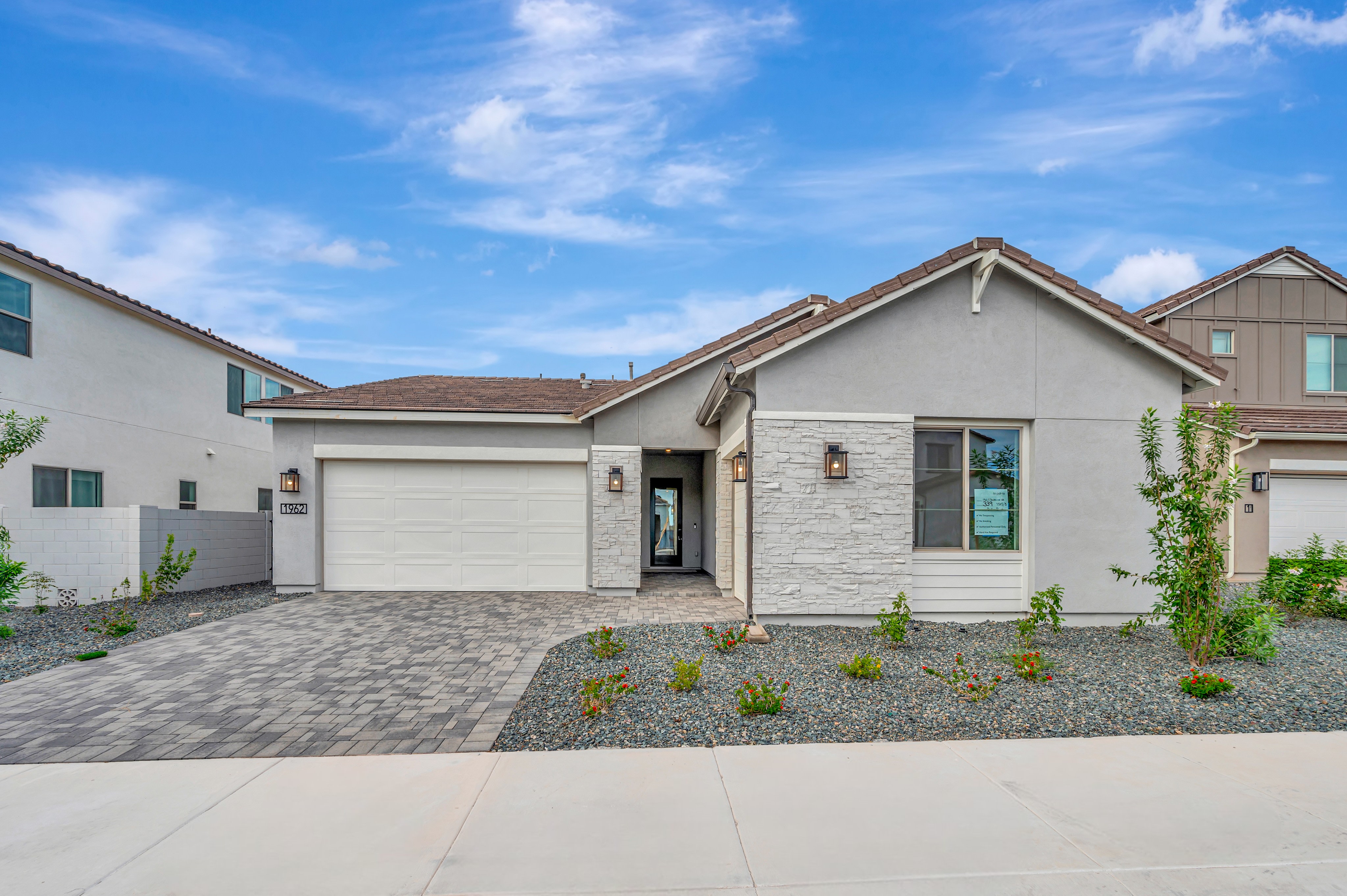 A modern single-story home with a white garage and stone accents sits behind a paved driveway and gravel landscaping under a bright blue sky.