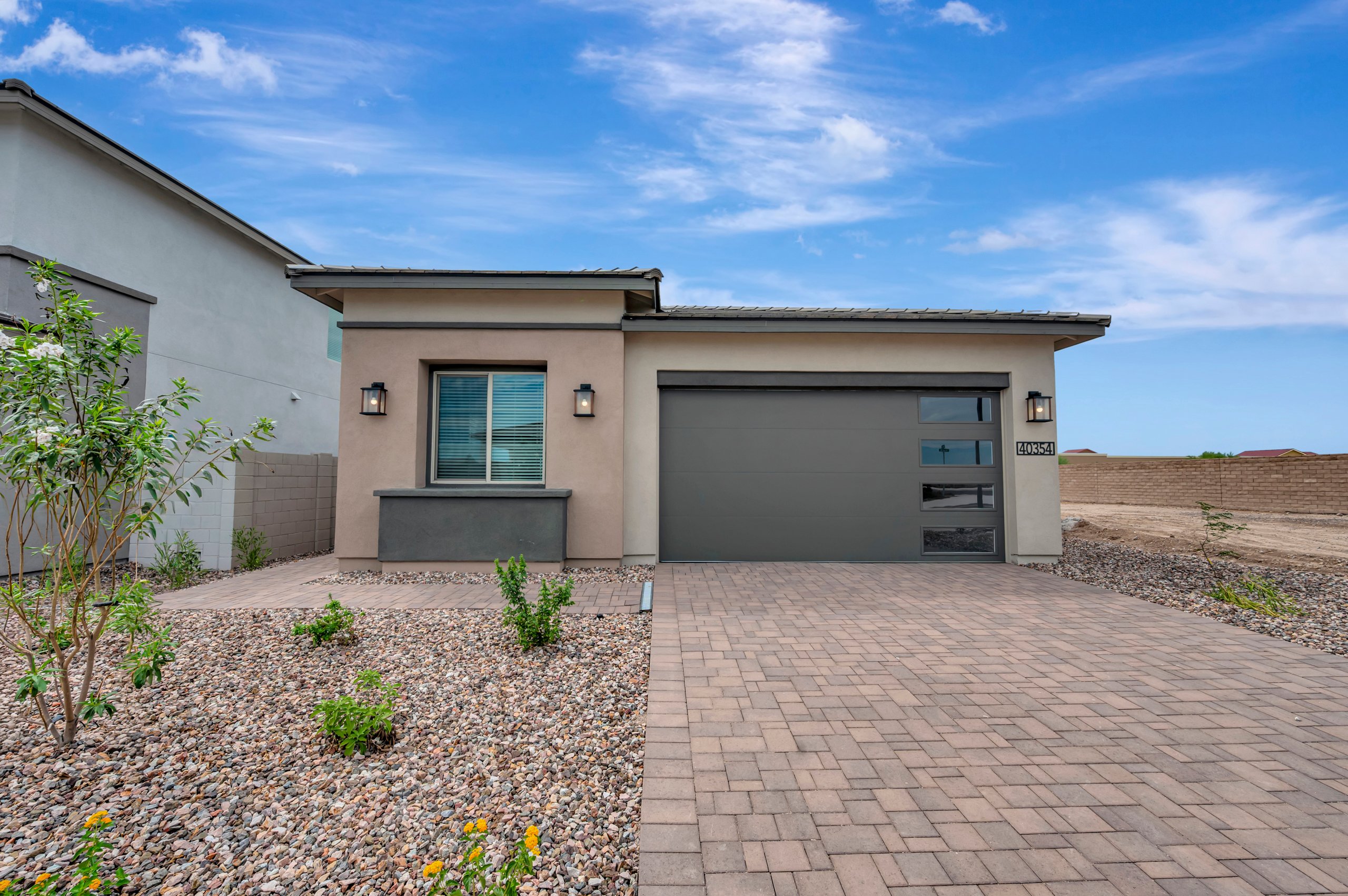 A modern single-story home with a grey garage door and paved driveway sits behind a gravel landscape under a bright blue sky.