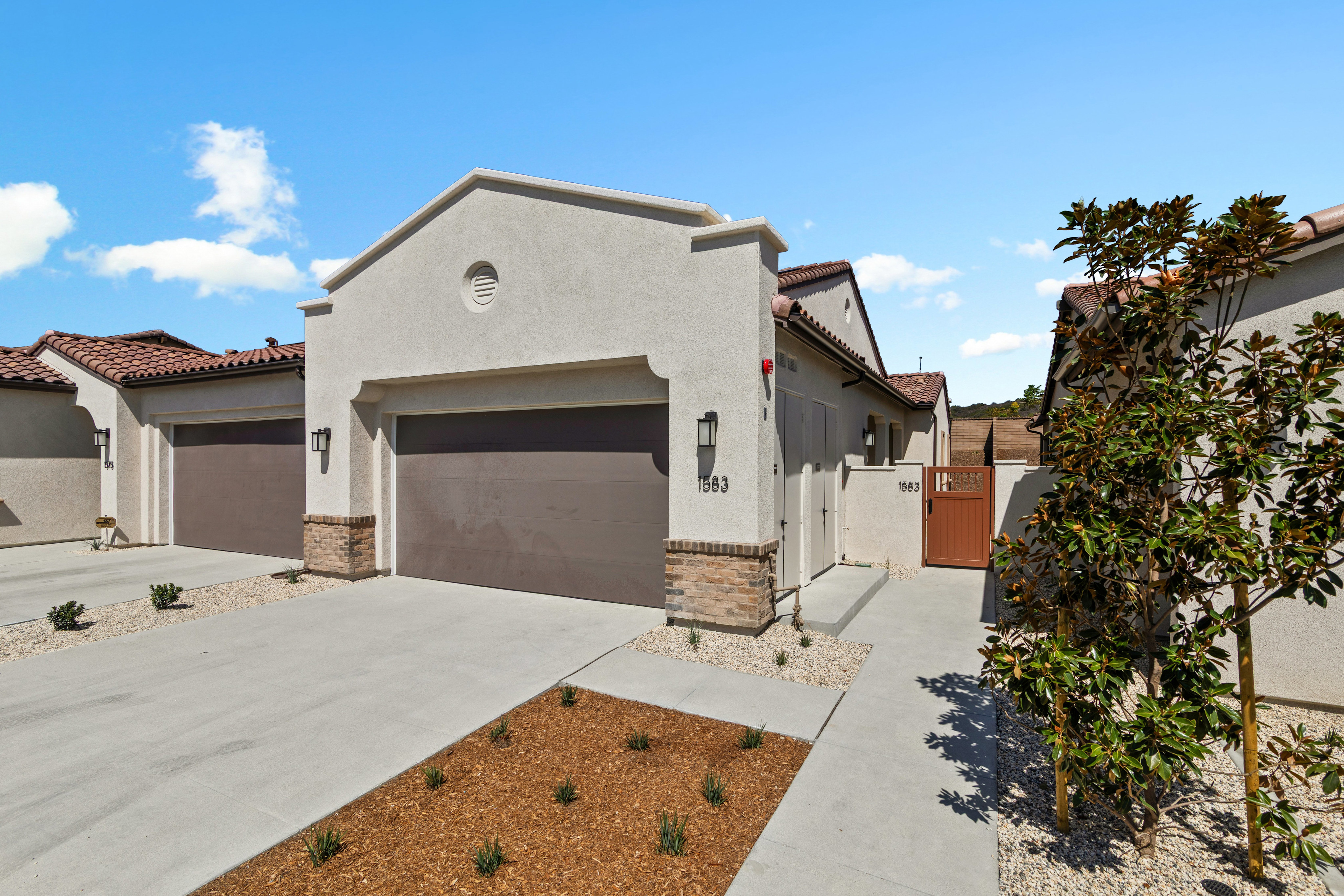A concrete driveway and small landscaped garden sit in the foreground of modern, light-colored stucco homes under a bright blue sky.