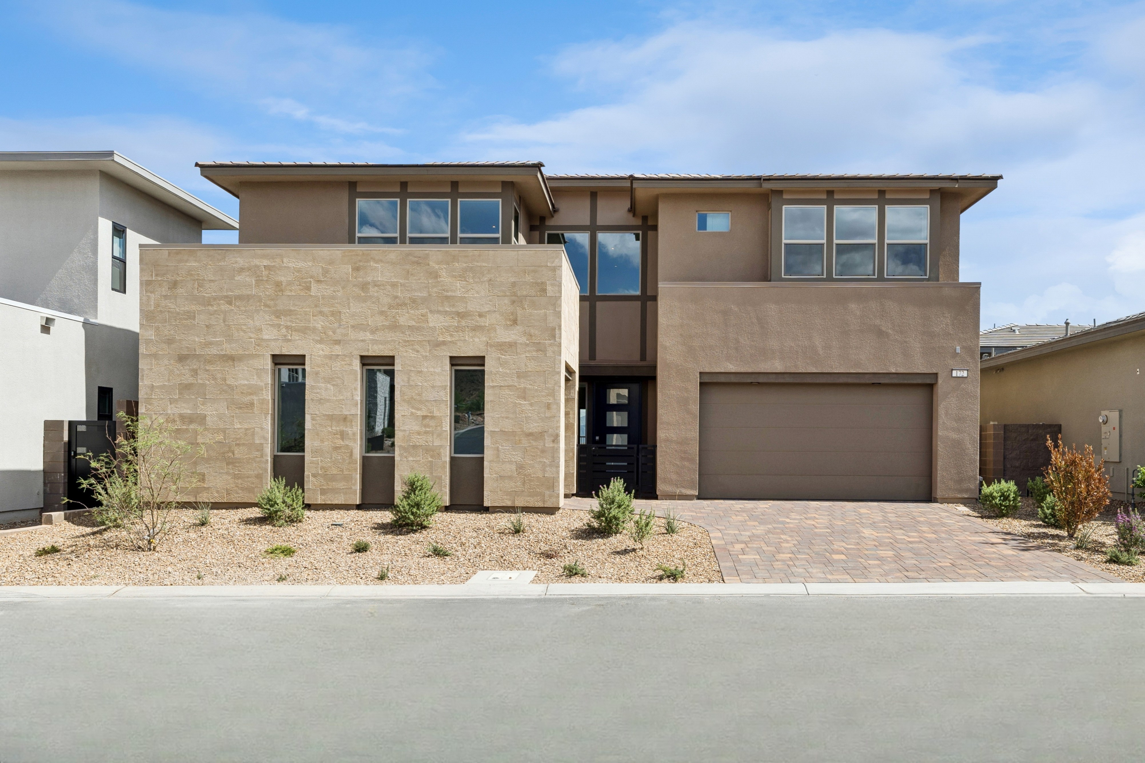 A modern two-story home with tan stone and stucco walls sits behind a gravel yard and paved driveway under a blue sky.