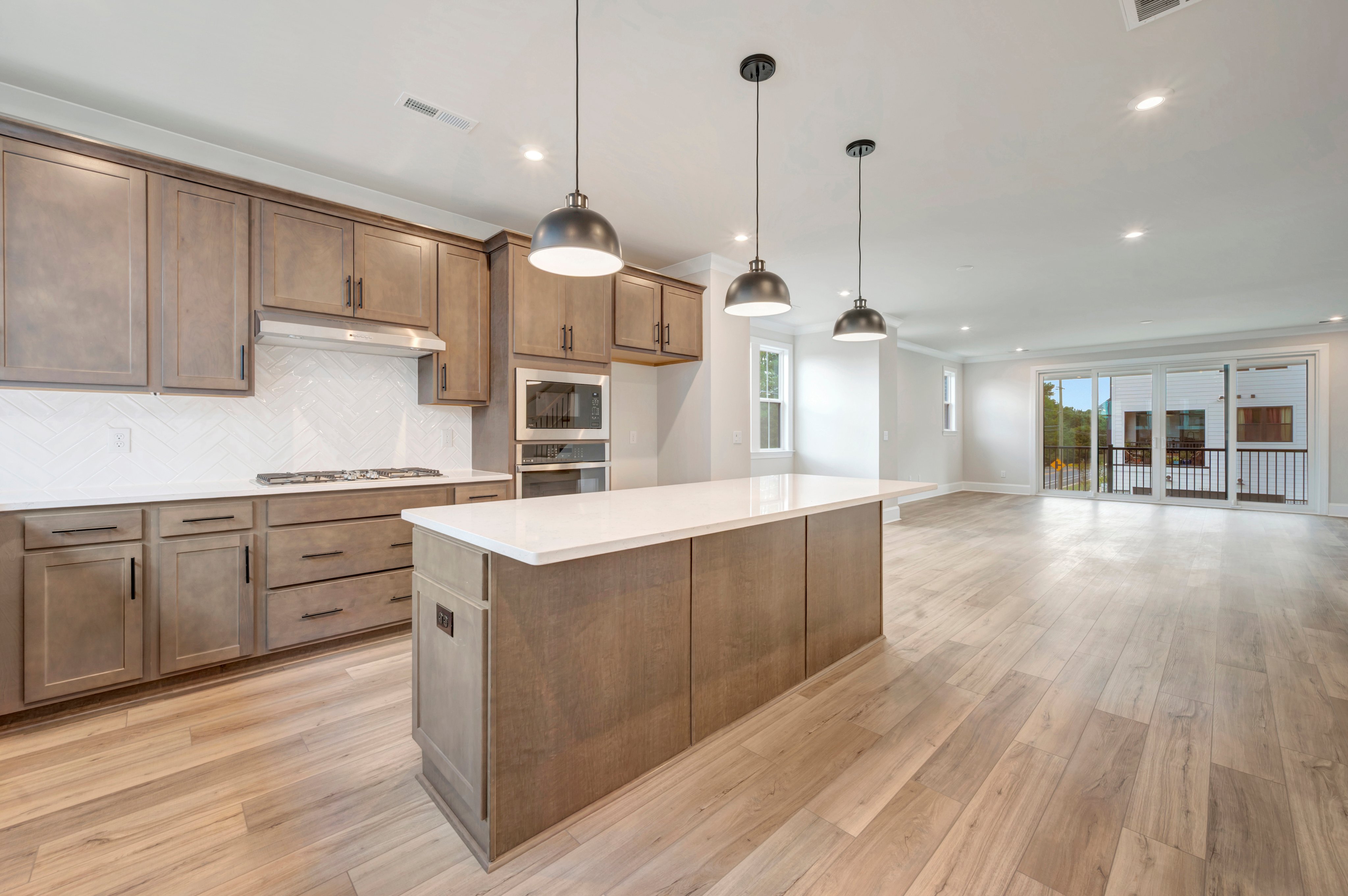 A modern kitchen with a large center island and grey cabinetry sits in the foreground, leading to an open living area with sliding glass doors in the background.