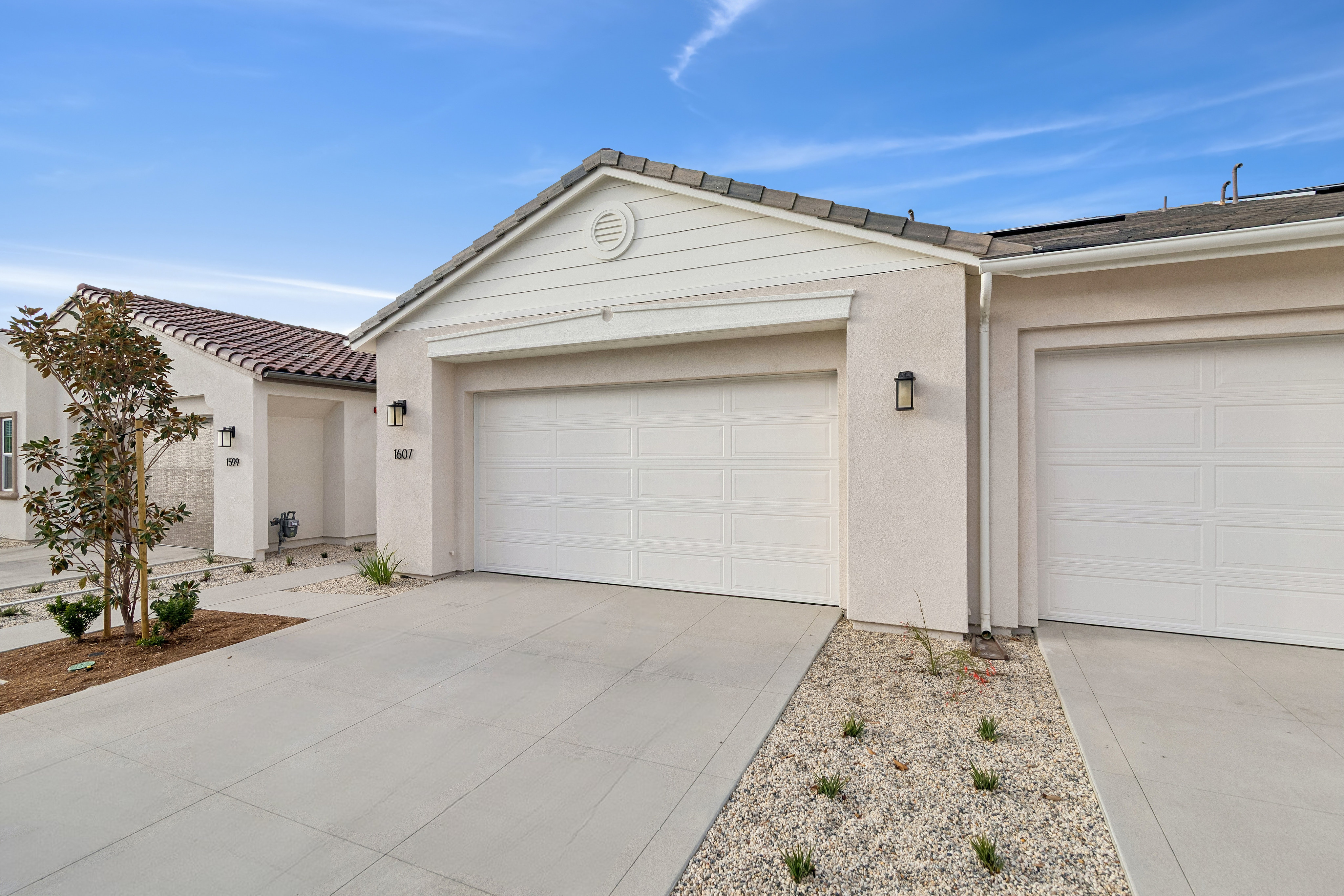 A concrete driveway and small landscaped garden sit in the foreground of two white garage doors under a clear blue sky.