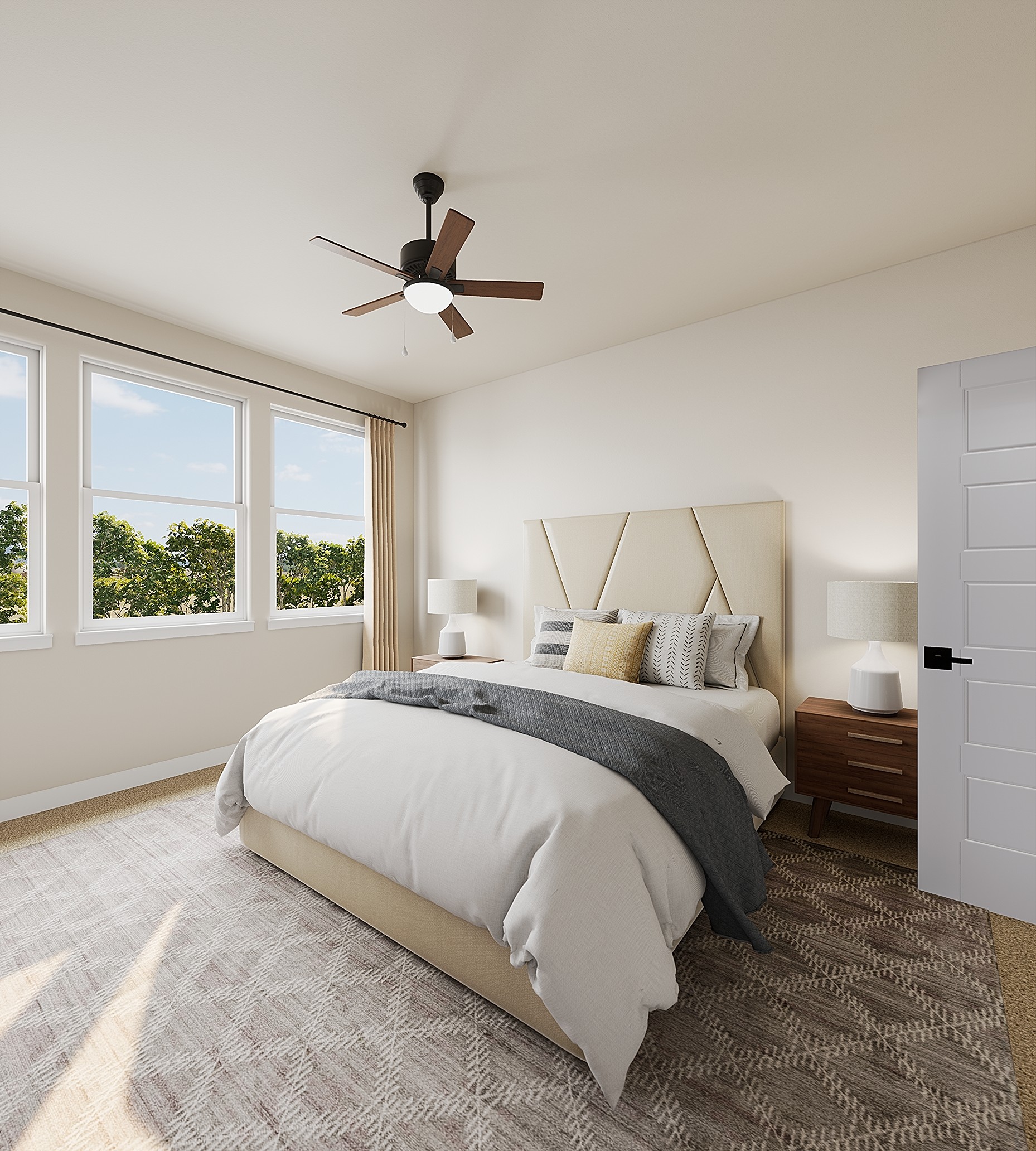 A neatly made bed with a grey throw sits on a patterned rug in a bright bedroom featuring a ceiling fan and large windows overlooking green trees.