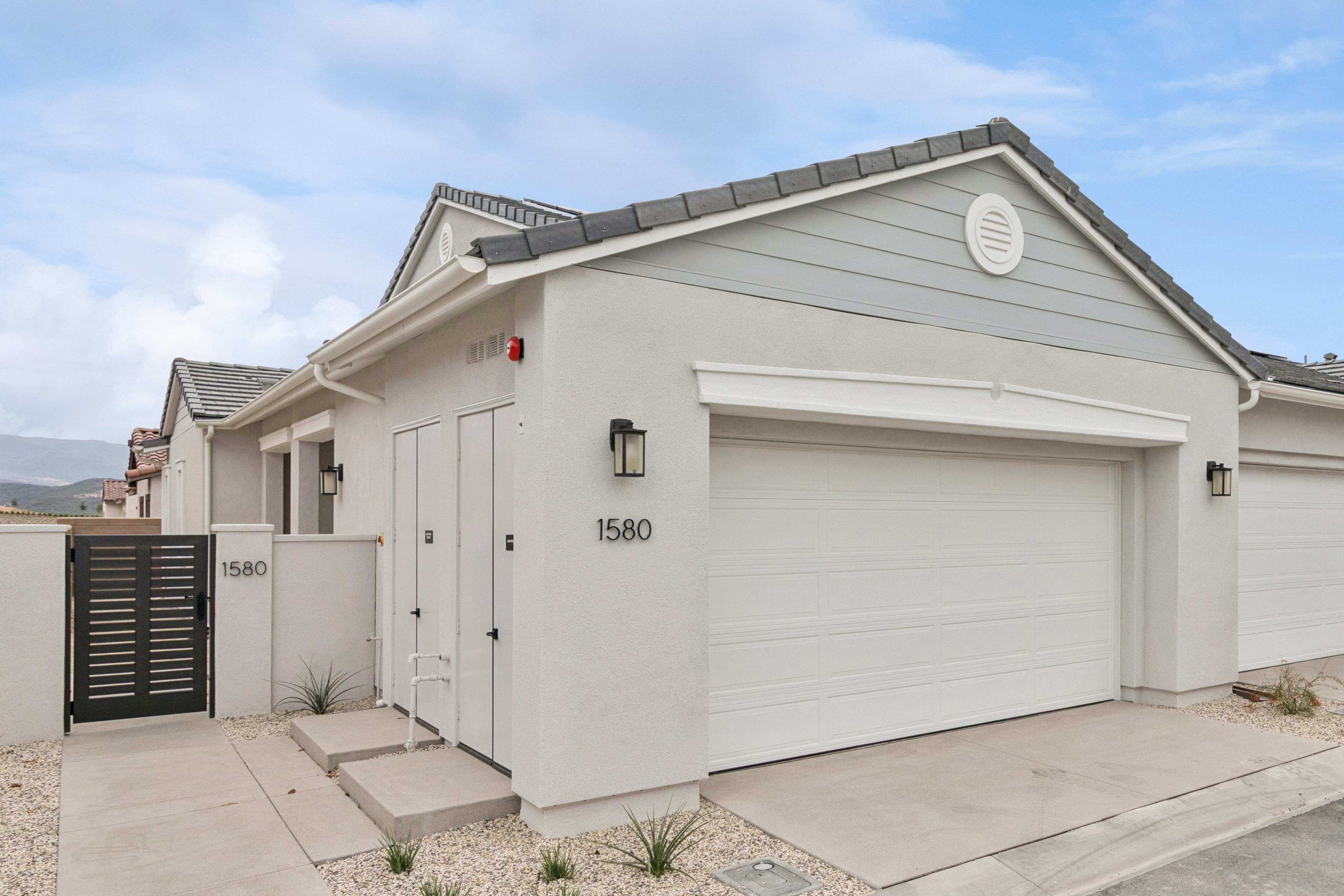 A modern light-gray house with a white garage door and black gate stands under a bright, cloudy blue sky.
