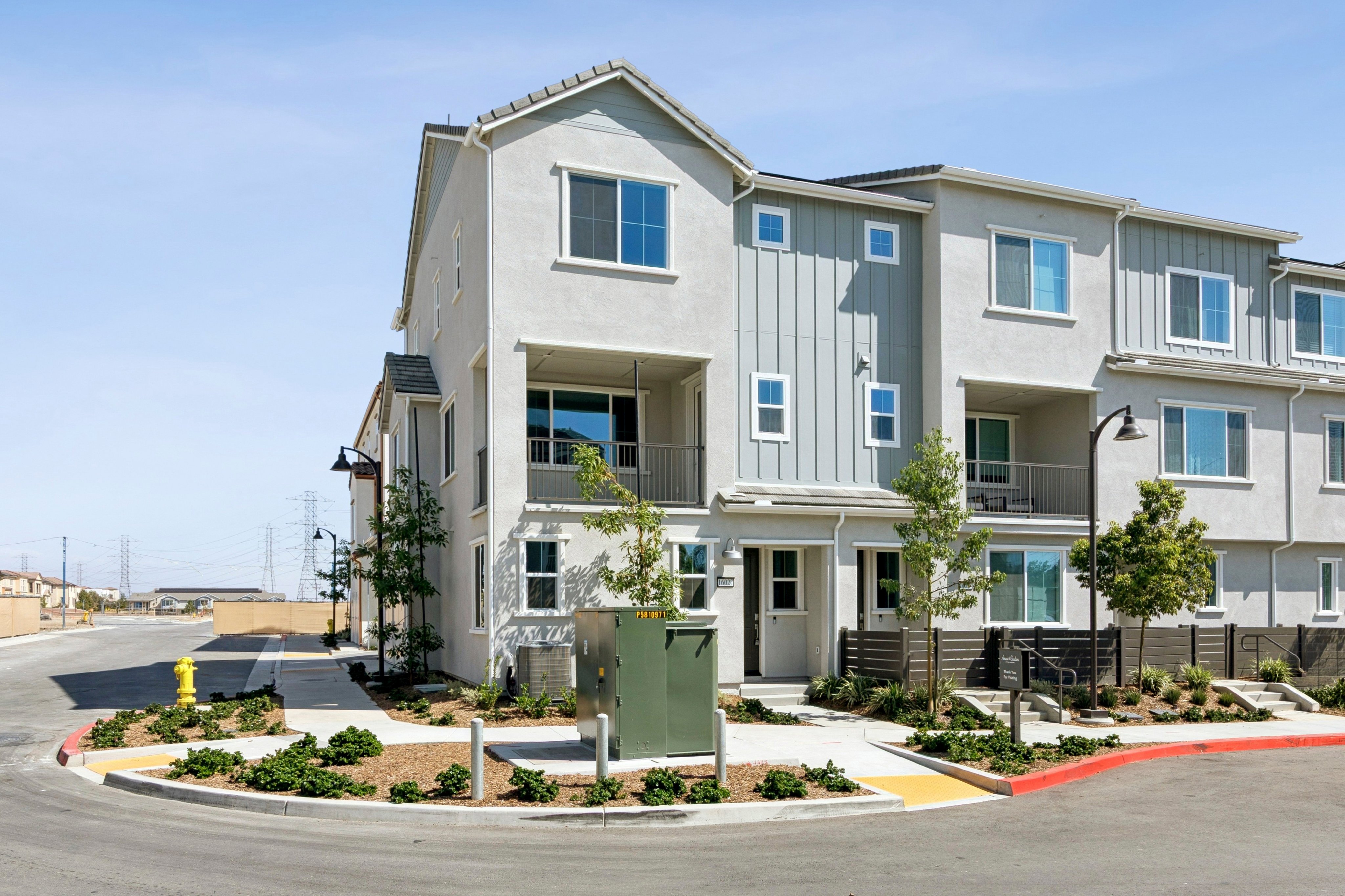 A modern multi-story apartment complex stands behind a paved street and landscaped foreground featuring a yellow fire hydrant and a green utility box.