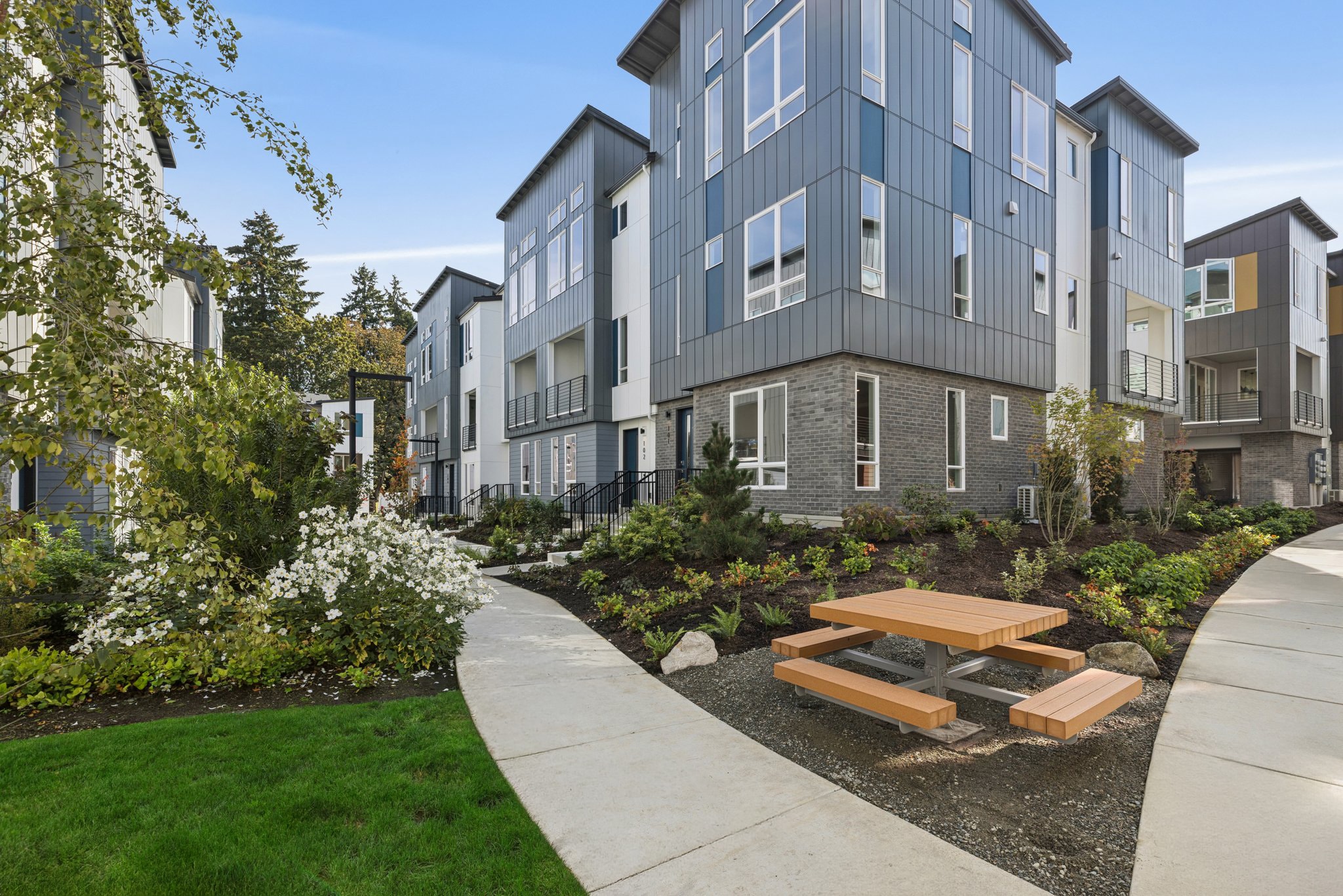 A wooden picnic table sits on a mulch bed beside a concrete walkway, with modern multi-story apartment buildings and lush greenery in the background.