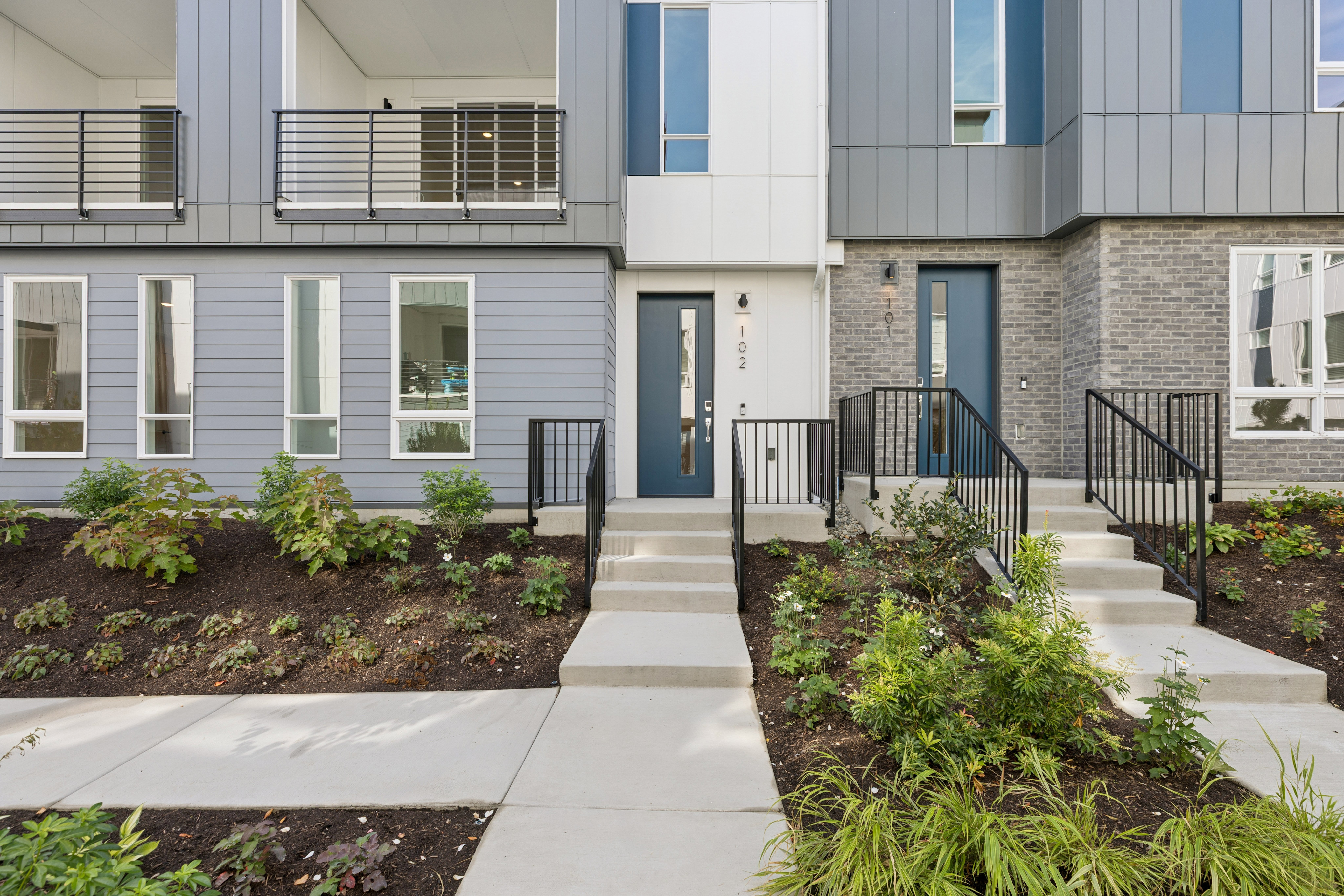 A concrete walkway and landscaped garden beds lead toward the entrances of a modern multi-story residential building.