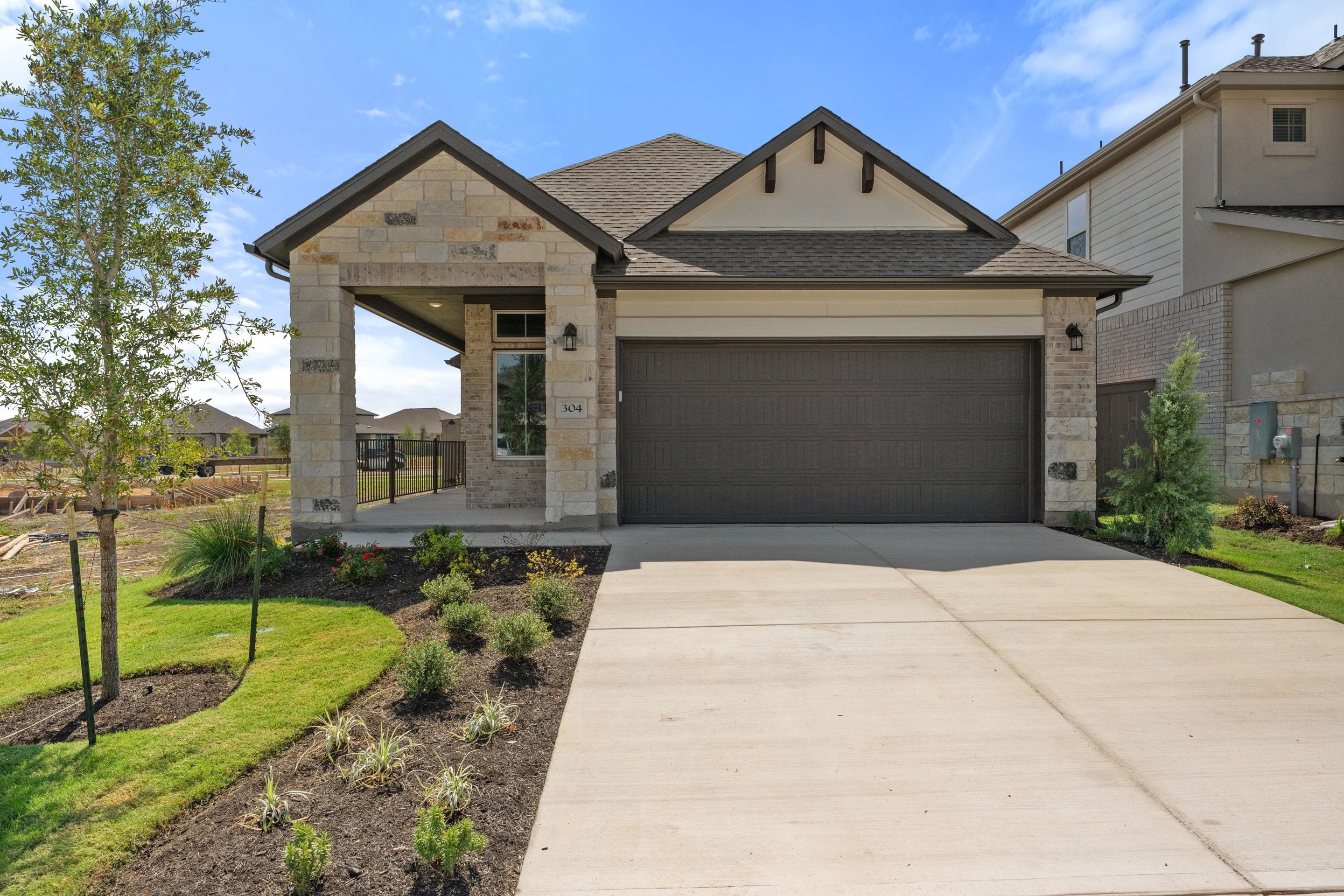 A modern stone and siding house with a dark brown garage door sits behind a concrete driveway and landscaped garden under a blue sky.