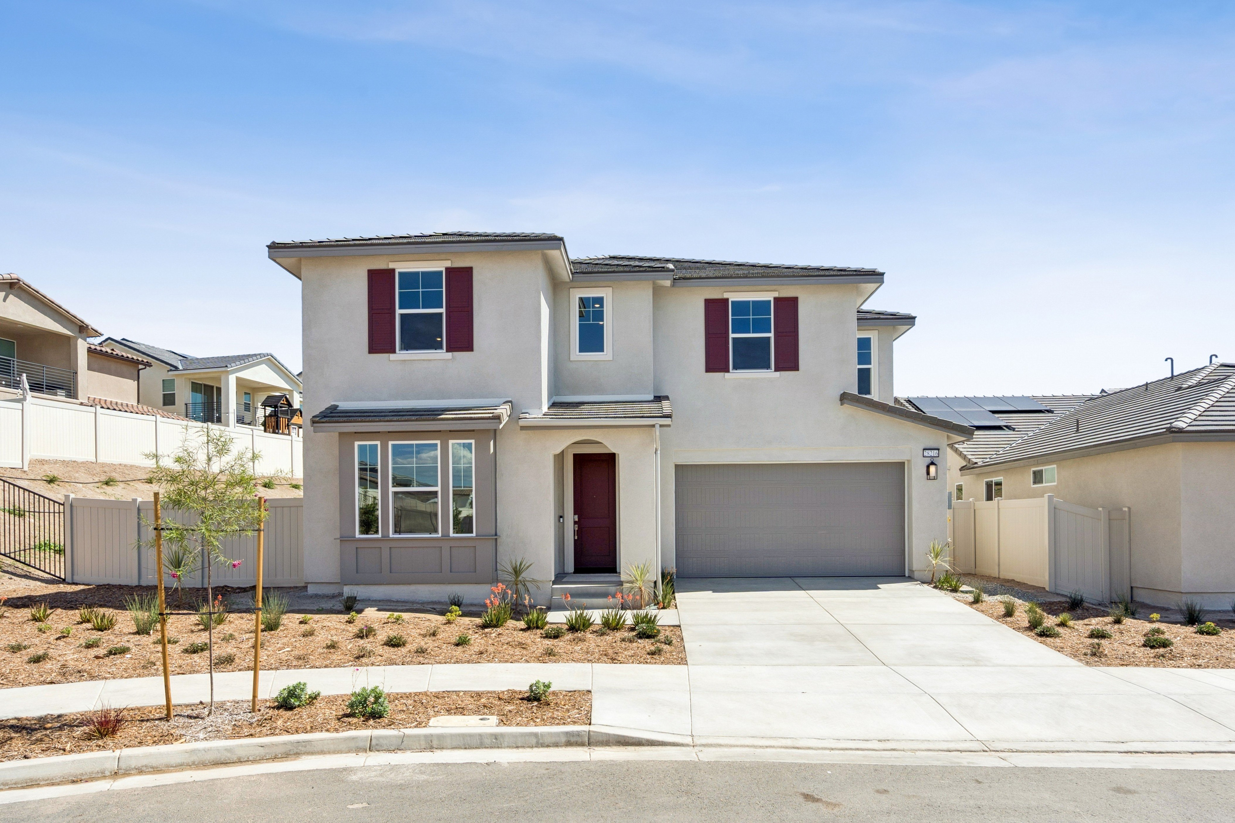 A two-story beige house with red shutters and a grey garage door sits behind a concrete driveway and small garden under a clear blue sky.