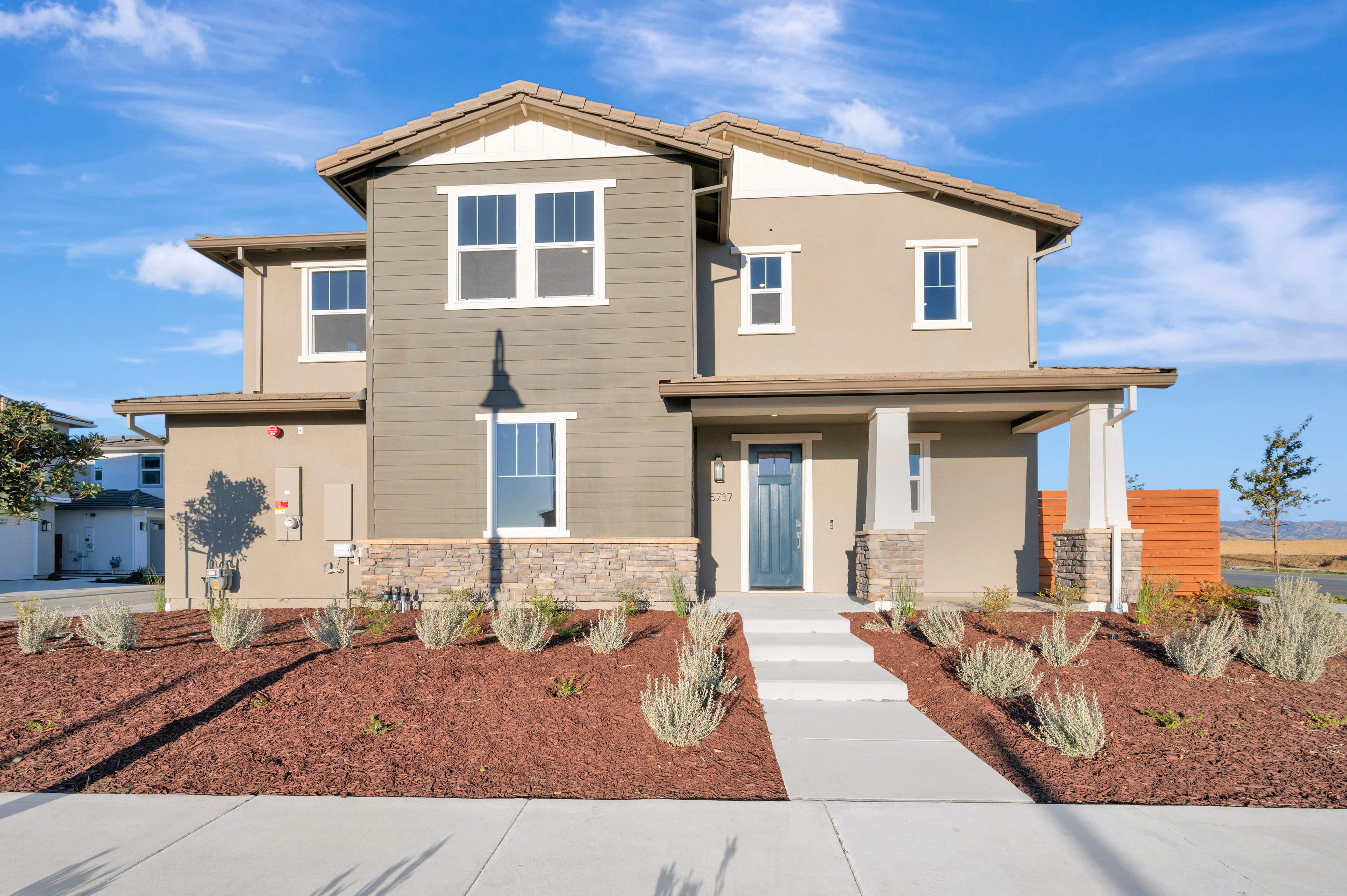 A modern two-story house with grey and beige siding stands behind a landscaped yard of red mulch and small shrubs under a bright blue sky.