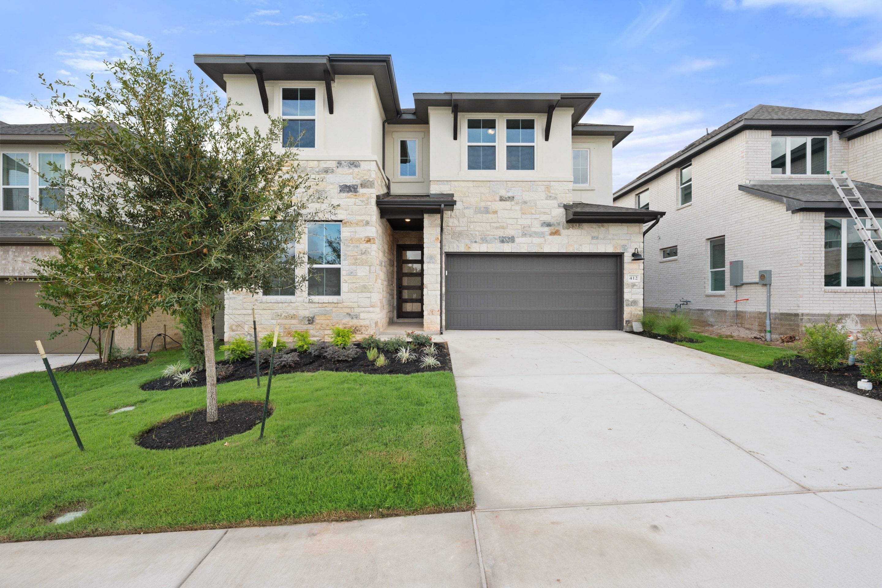 A modern two-story stone and stucco home with a grey garage door stands behind a green lawn and a small tree under a blue sky.