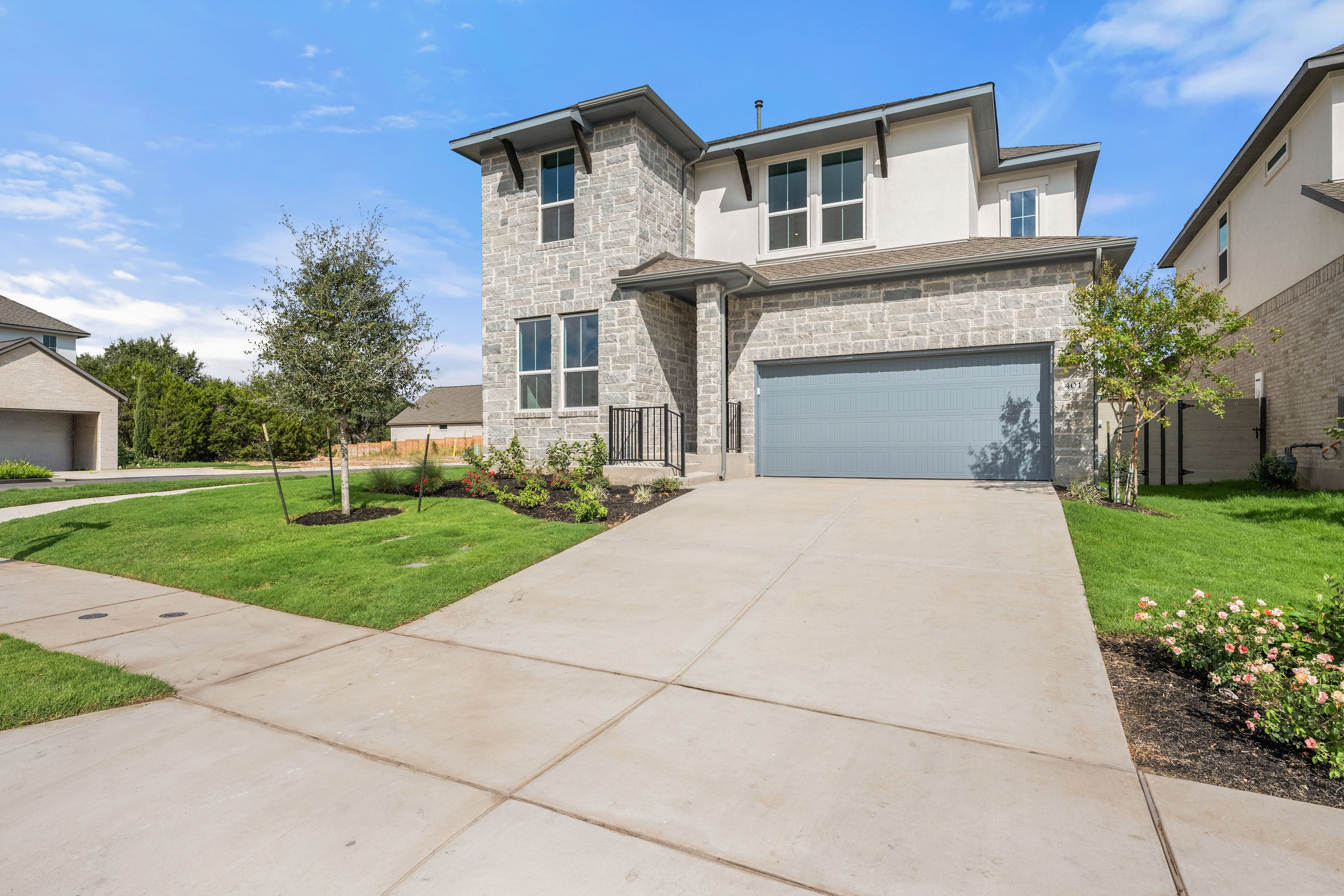 A concrete driveway and green lawn lead to a modern two-story stone and stucco house under a bright blue sky.