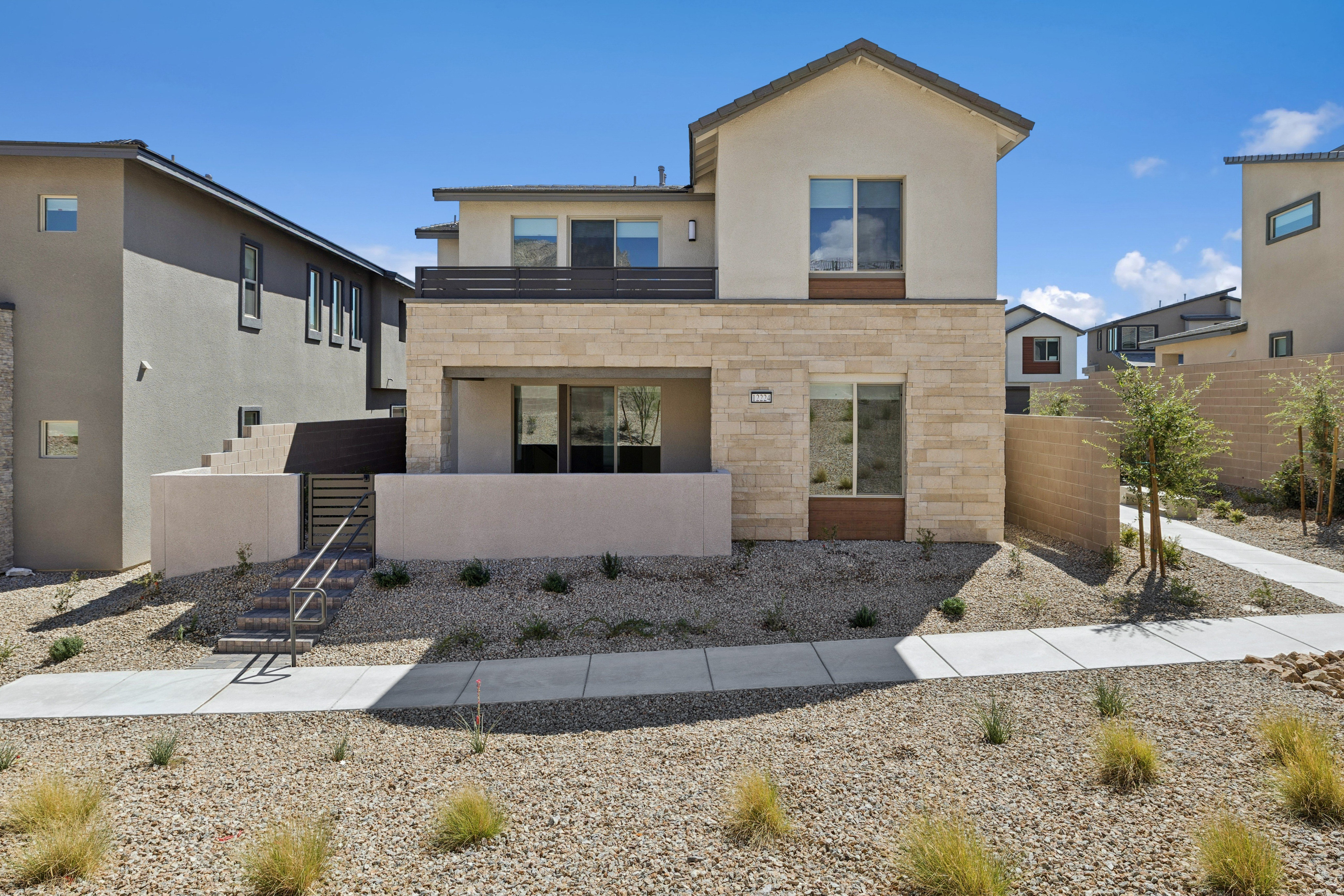 A modern two-story house with tan and beige siding stands behind a gravel yard and concrete walkway under a clear blue sky.