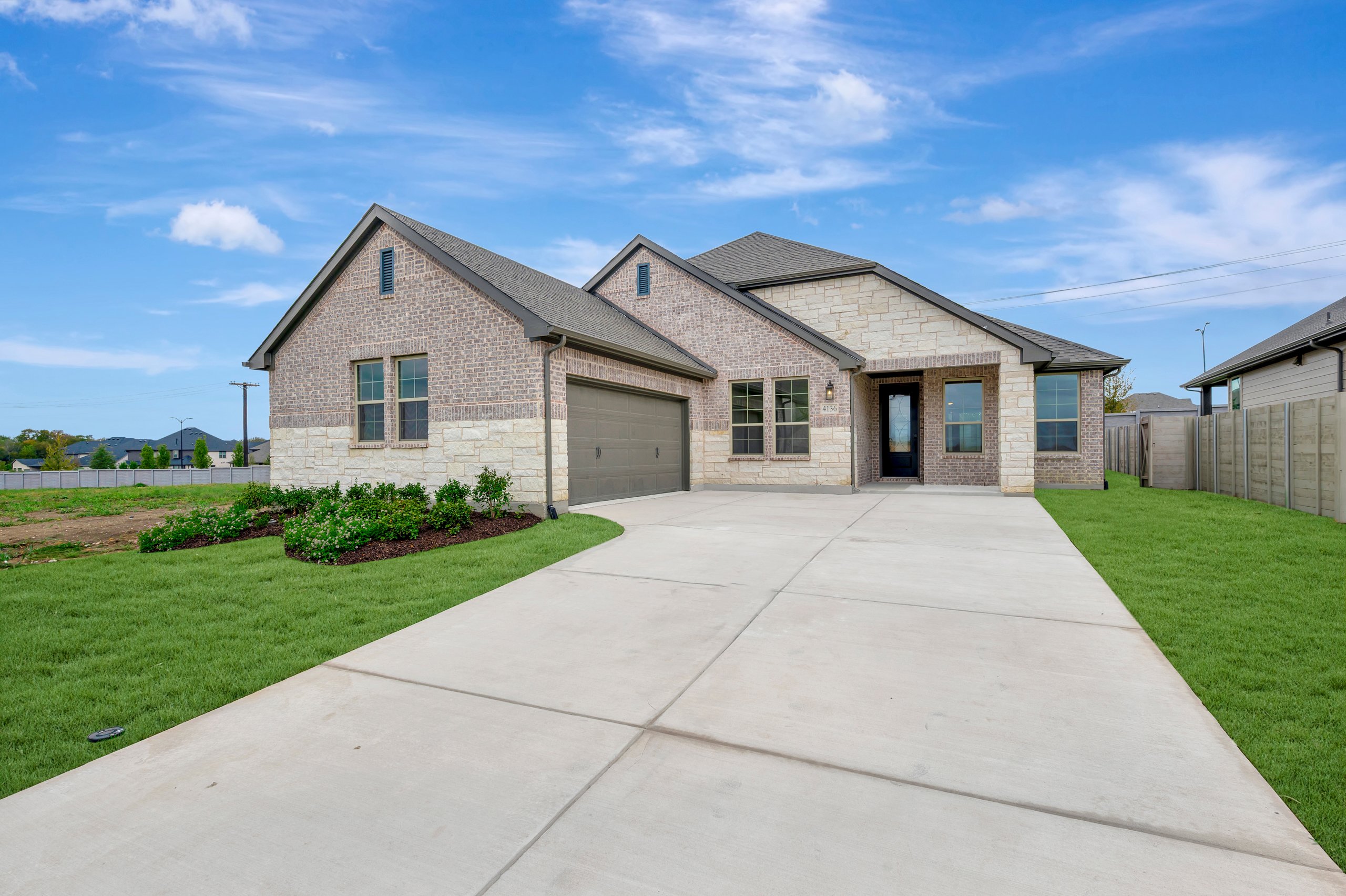 A wide concrete driveway leads to a modern brick and stone house set against a bright blue sky with wispy clouds.