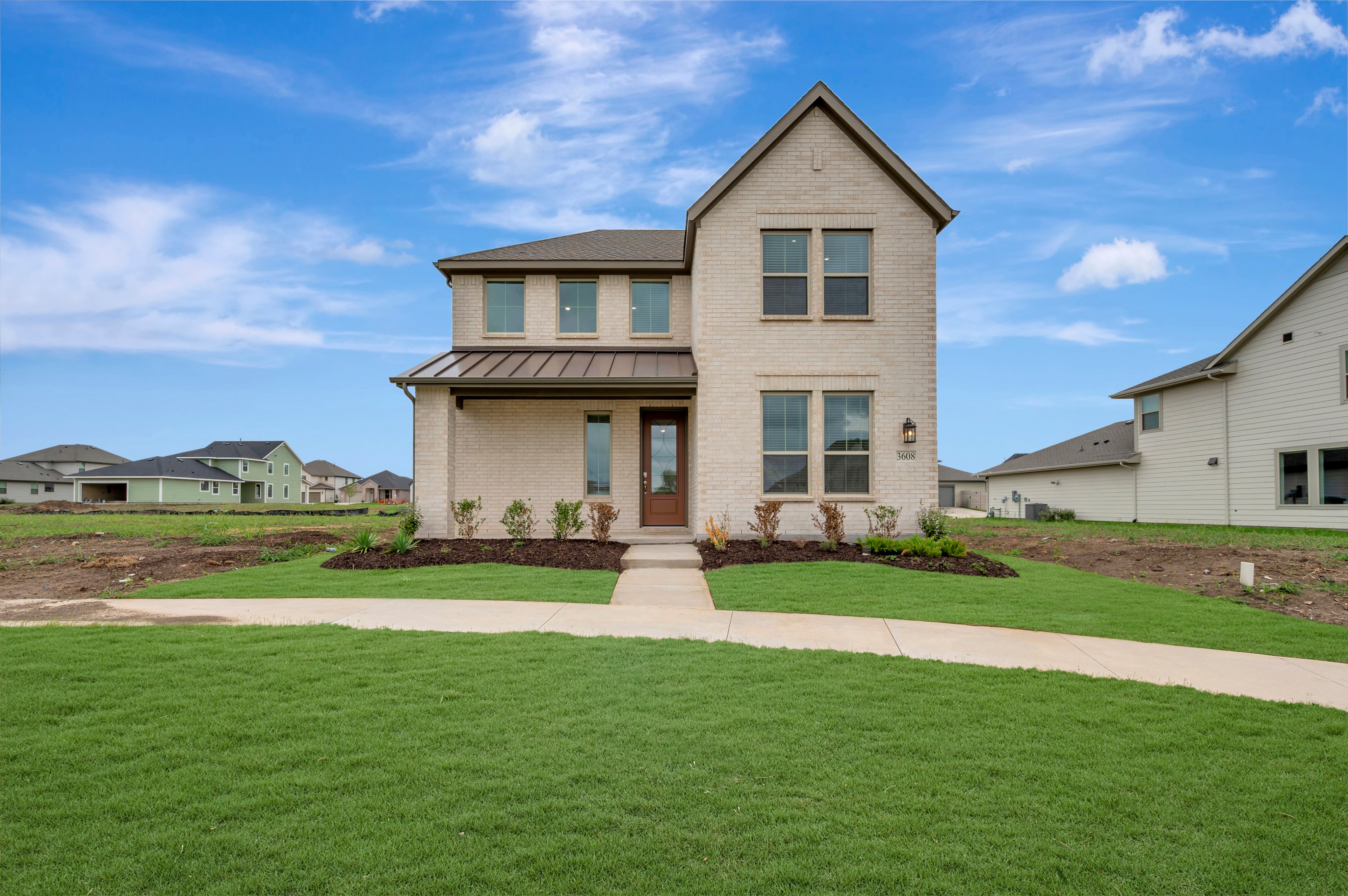 A two-story light-colored brick house stands behind a manicured green lawn and sidewalk under a bright blue sky with scattered clouds.