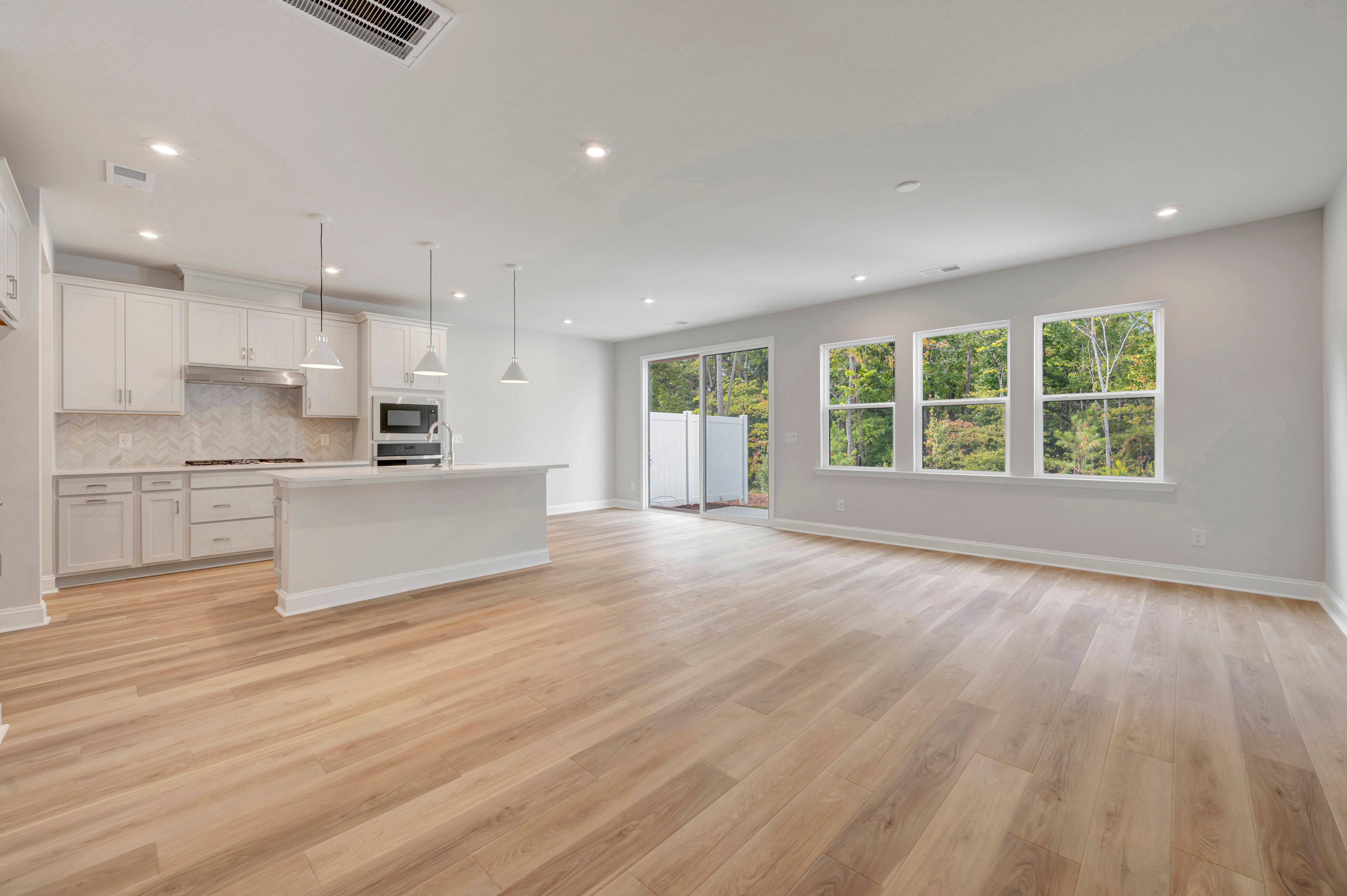 Light-colored hardwood floors fill the foreground of an open-concept living space, leading back to a modern white kitchen and large windows overlooking a wooded area.