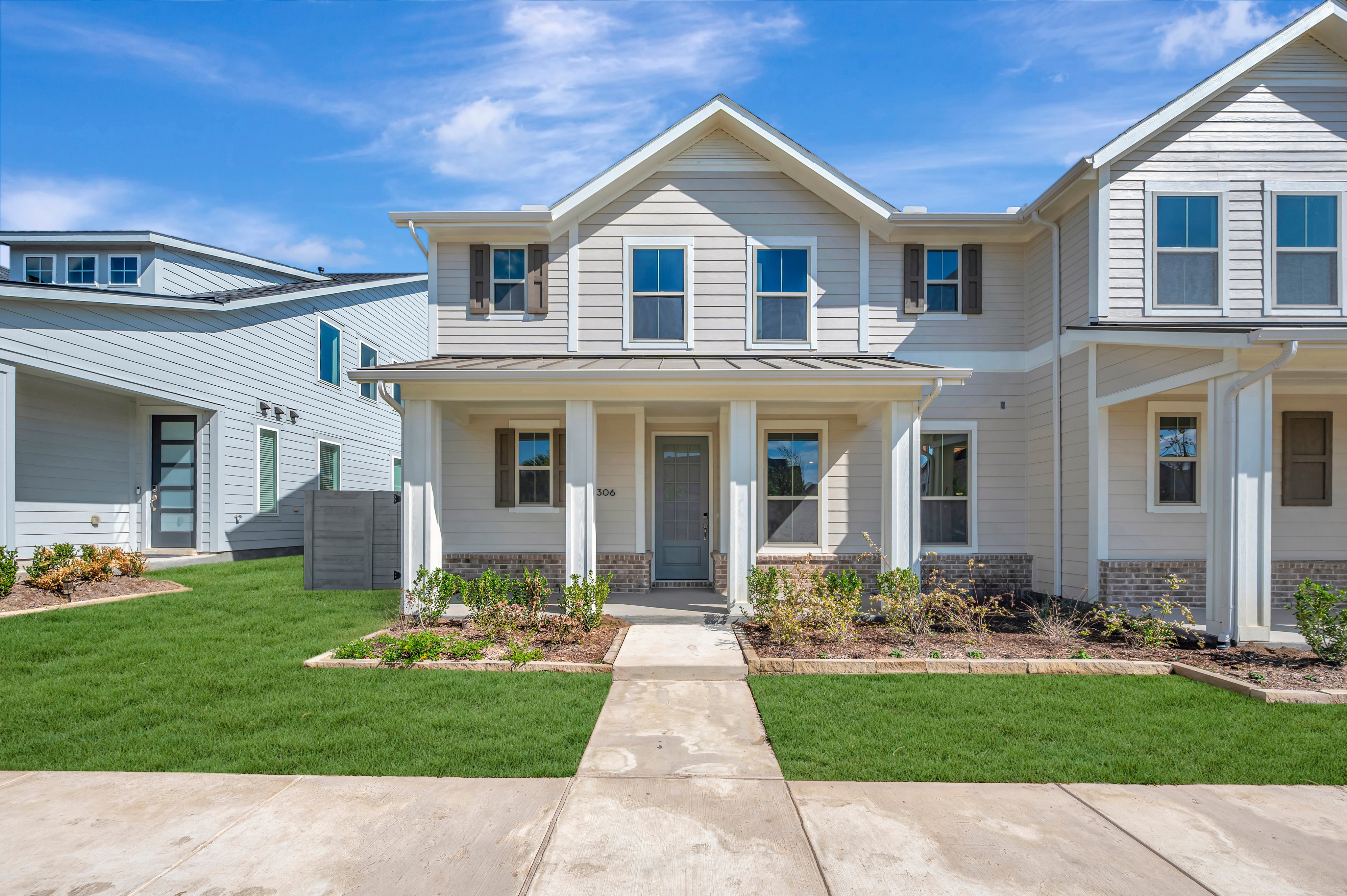 A light-colored two-story house with a front porch and small garden beds sits behind a concrete sidewalk under a bright blue sky.