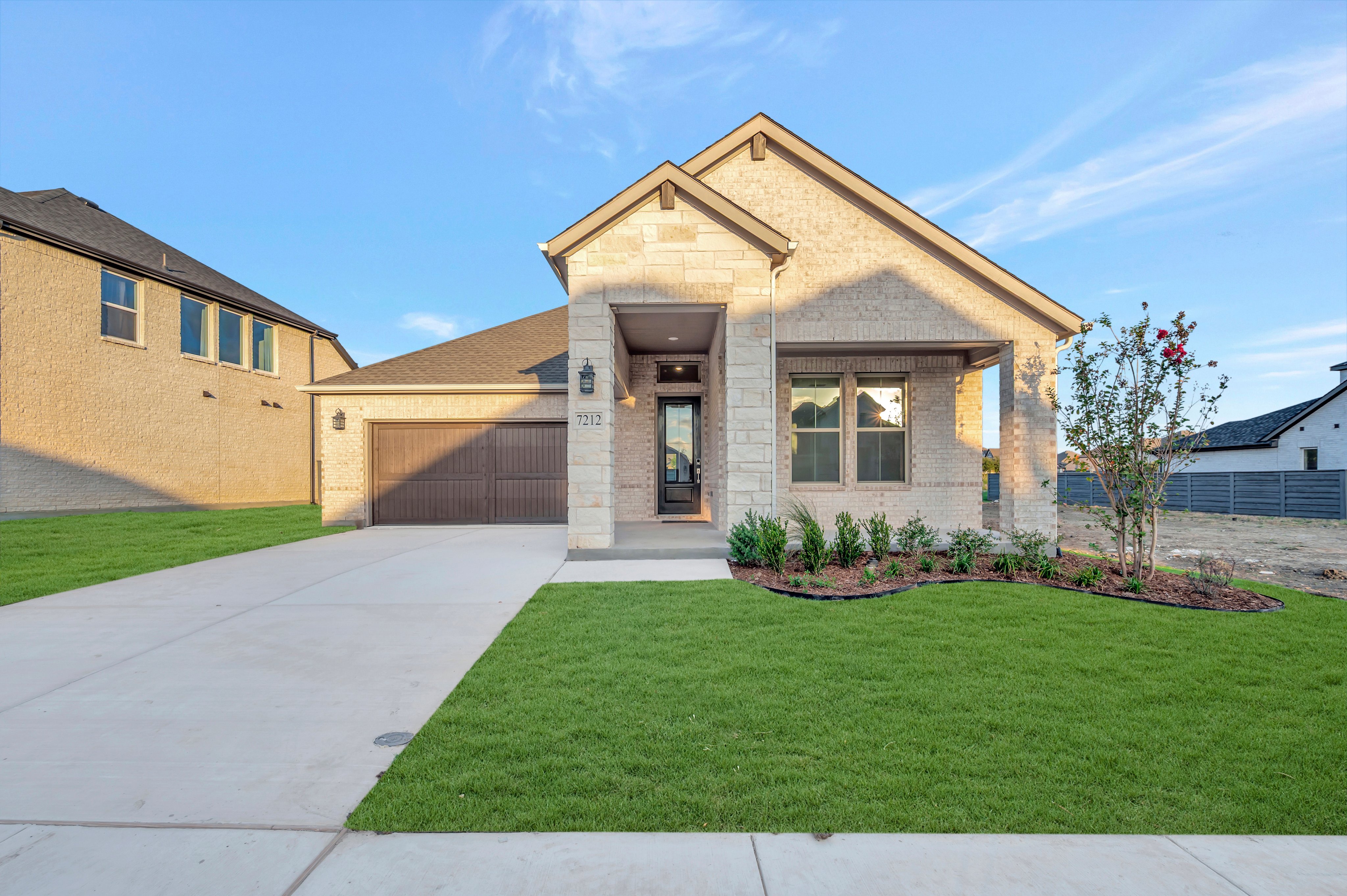 A modern light-colored stone house with a brown garage door sits behind a green lawn and concrete driveway under a clear blue sky.