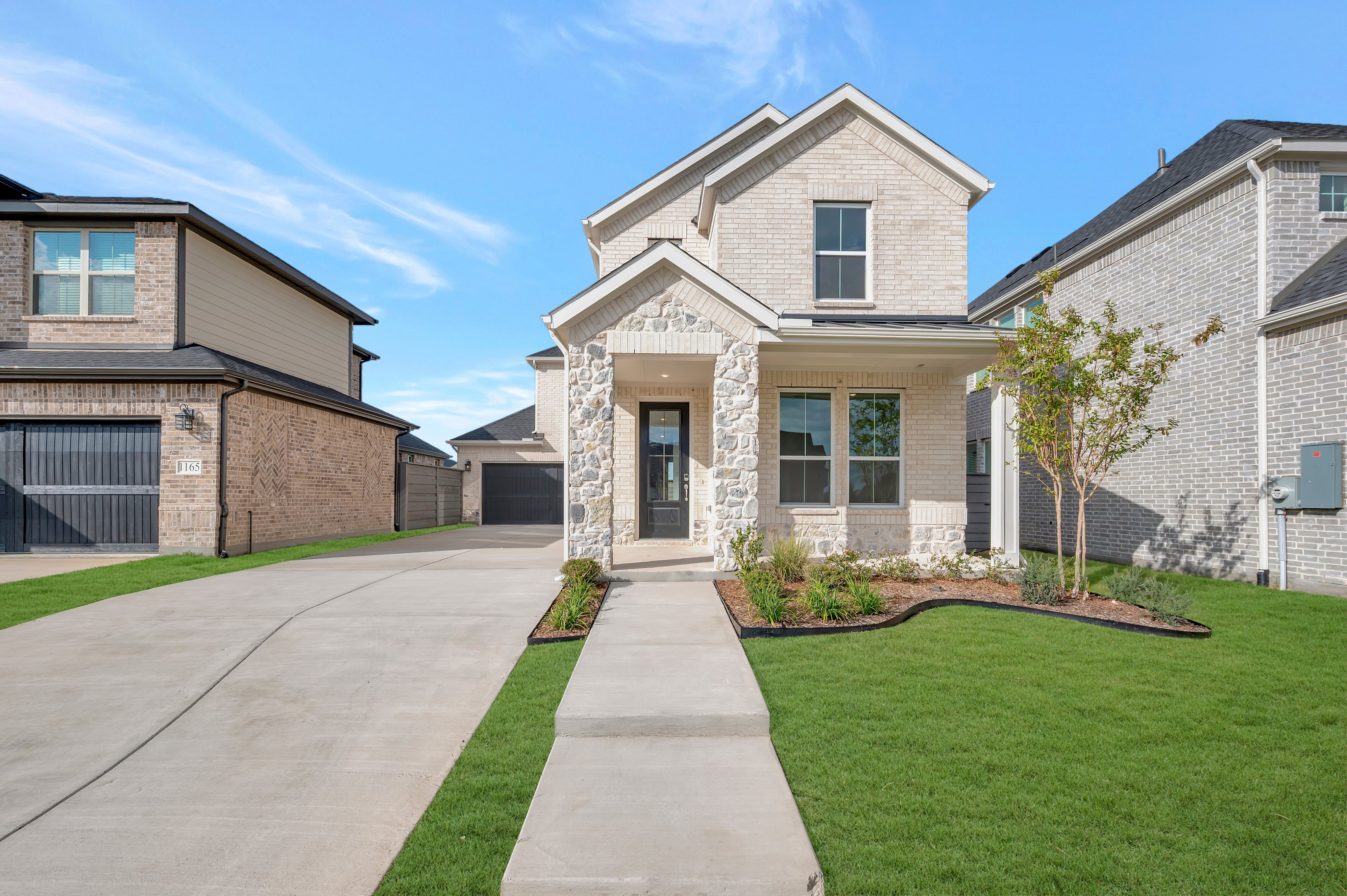 A concrete driveway and walkway lead to a modern two-story stone house, flanked by neighboring homes under a clear blue sky.