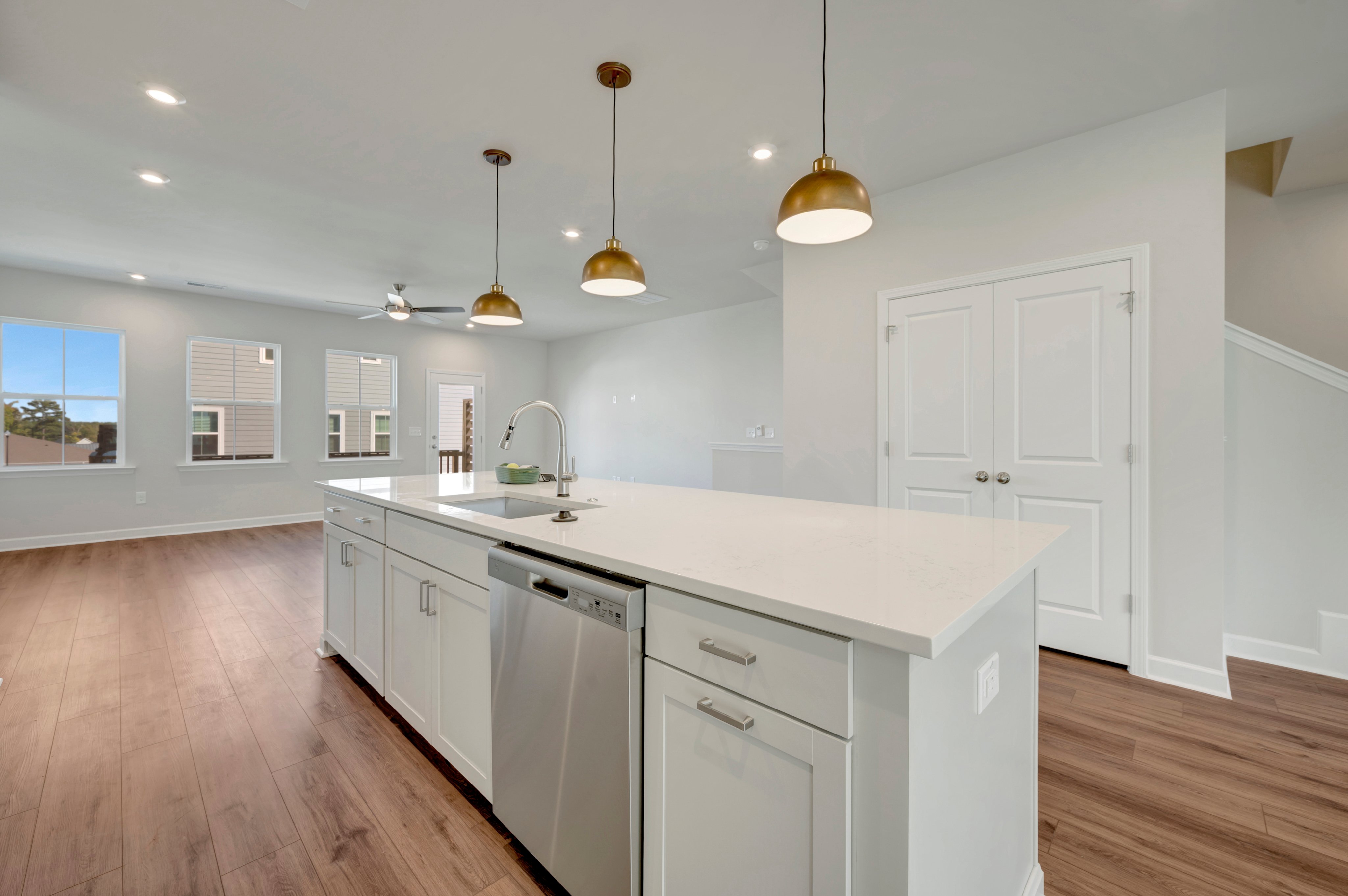 A modern white kitchen island with a stainless steel dishwasher sits in the foreground of a bright, open room with hardwood floors and white double doors in the background.