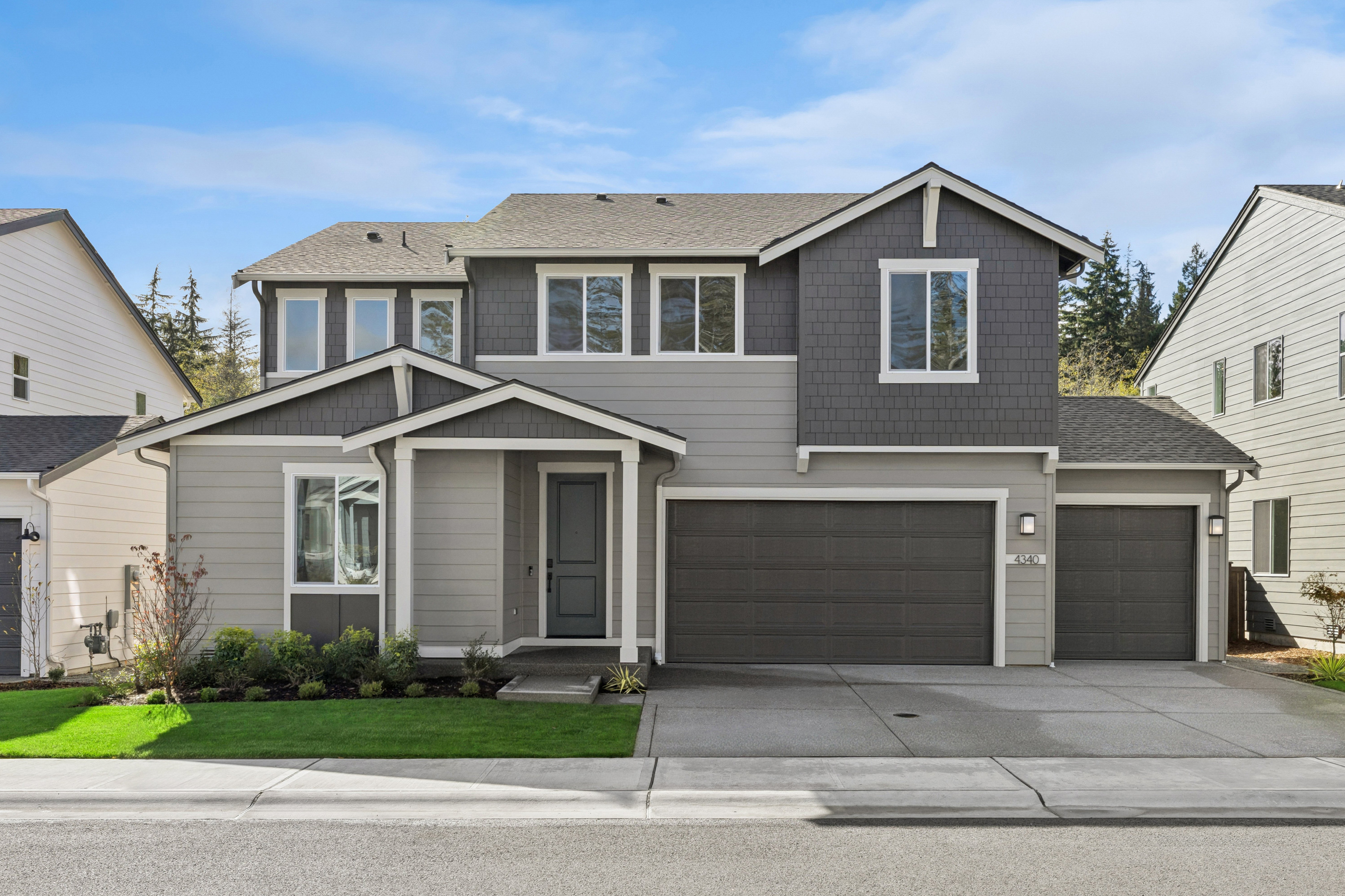 A modern two-story grey house with a concrete driveway and green lawn sits under a clear blue sky.