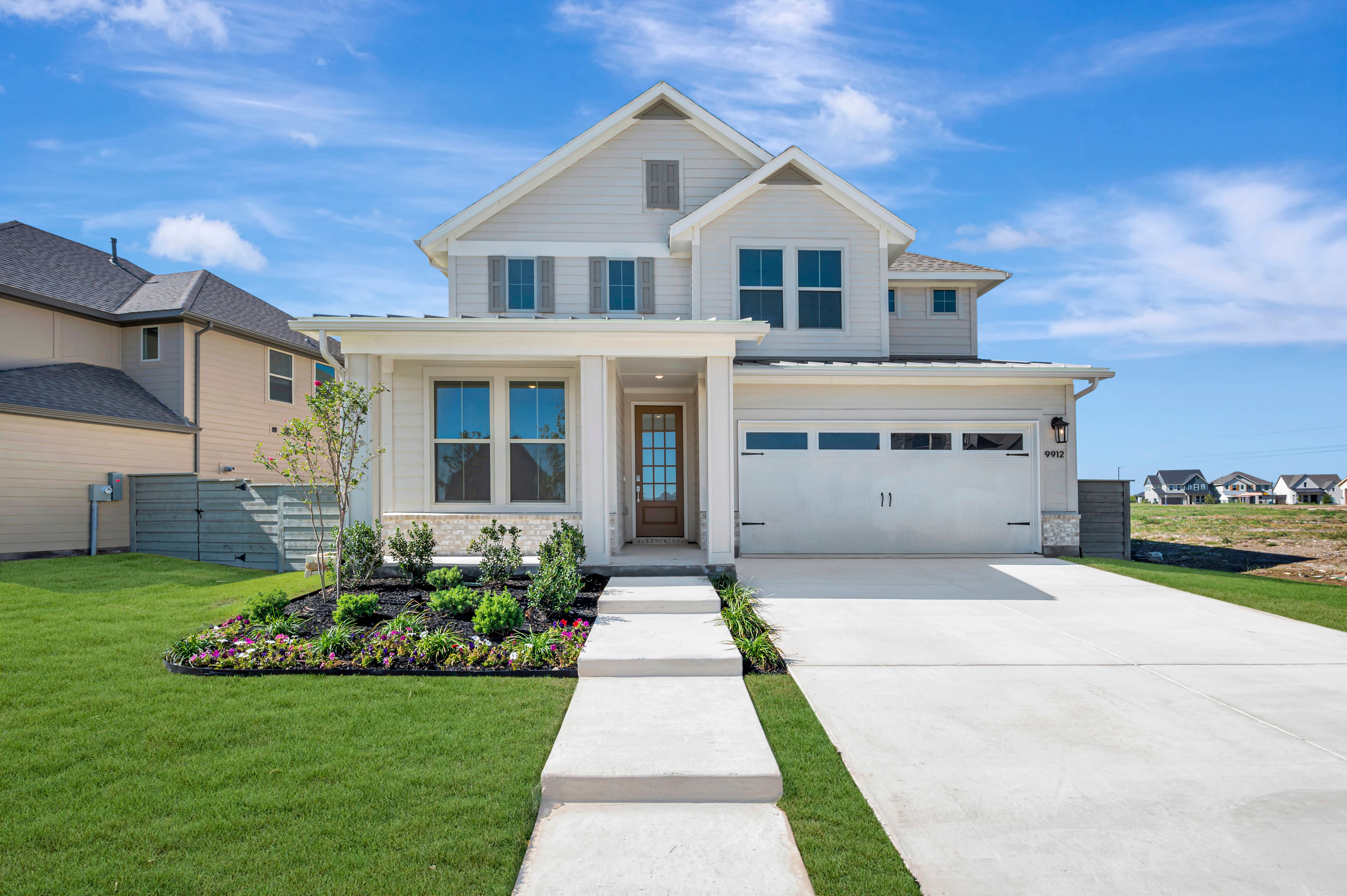 A modern two-story white house with a concrete driveway and manicured lawn sits under a bright blue sky with scattered clouds.