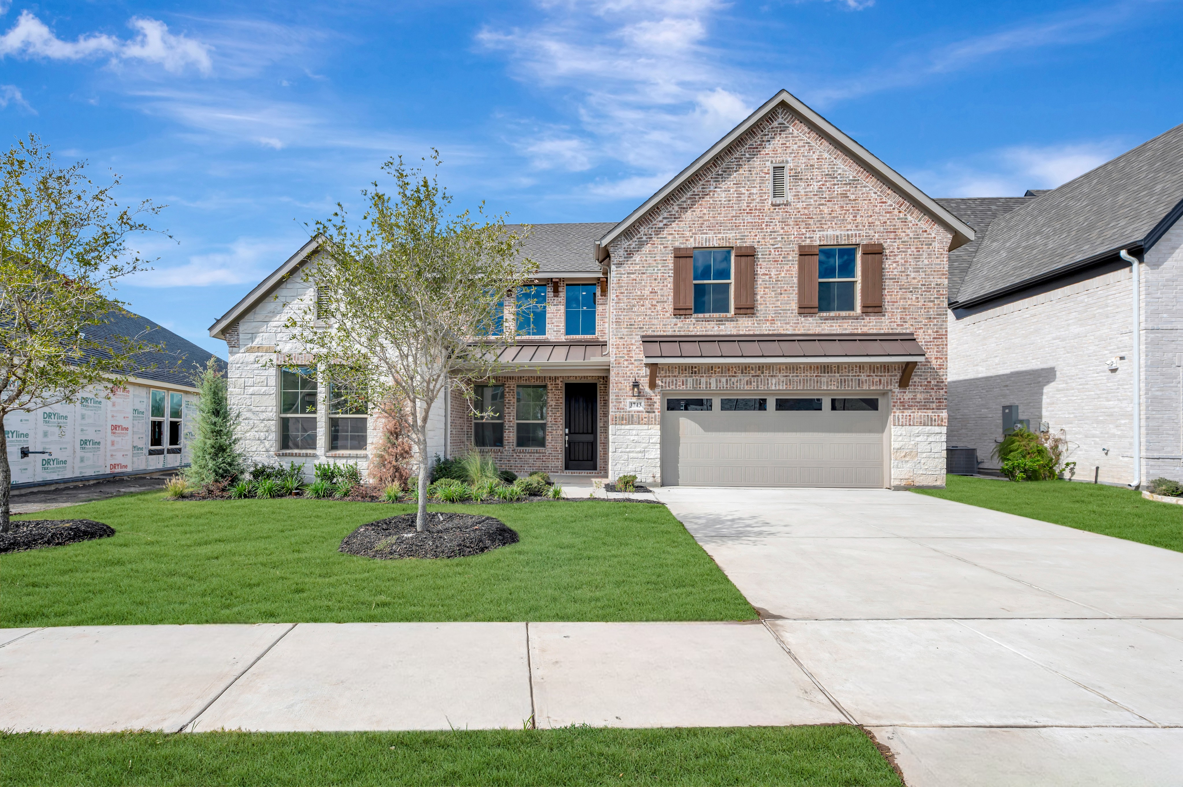 A large brick and stone house with a concrete driveway and green lawn sits under a bright blue sky with wispy clouds.
